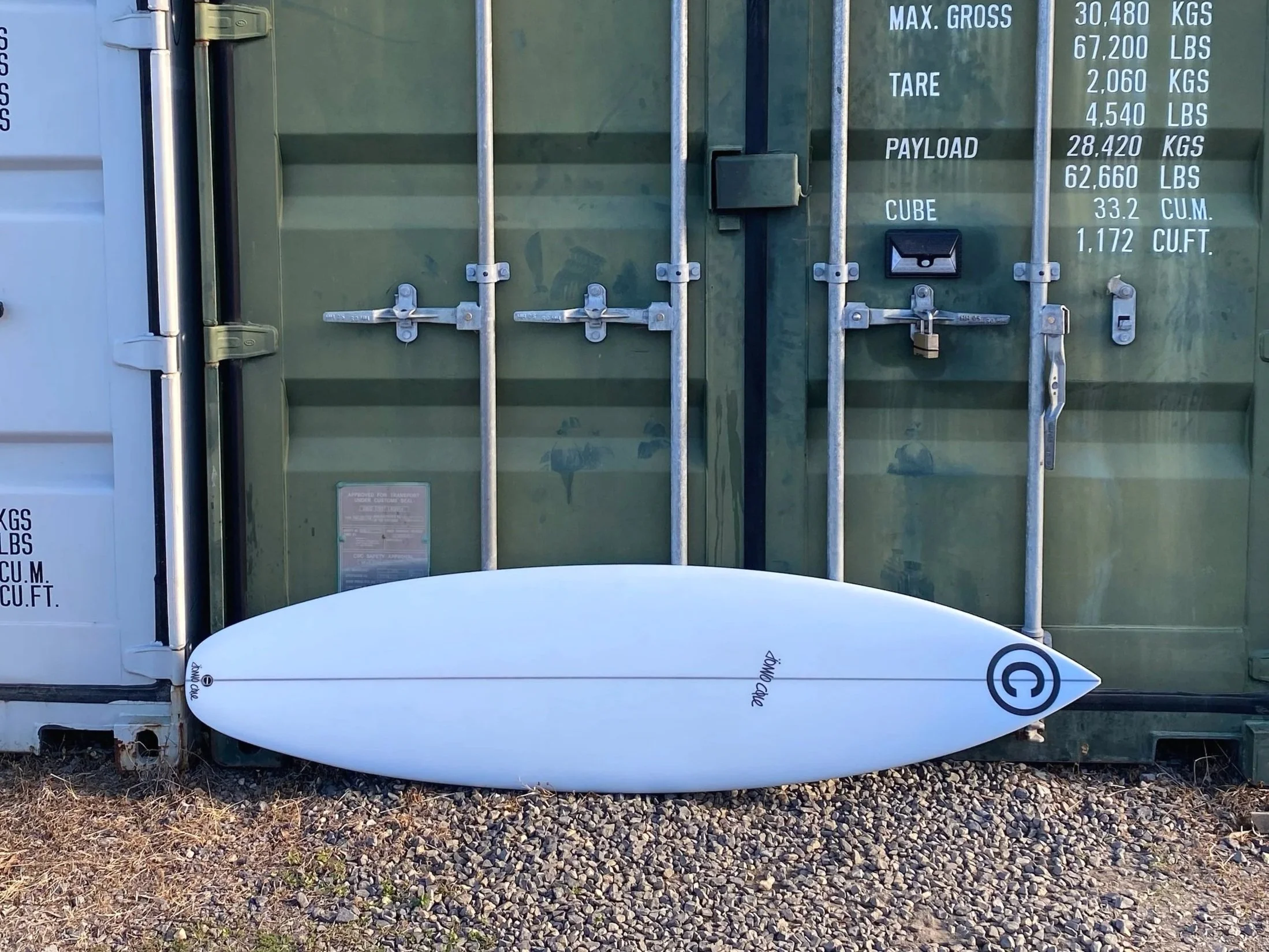 White surfboard with black logo and text resting against a shipping container with metal locking bars and weight information.