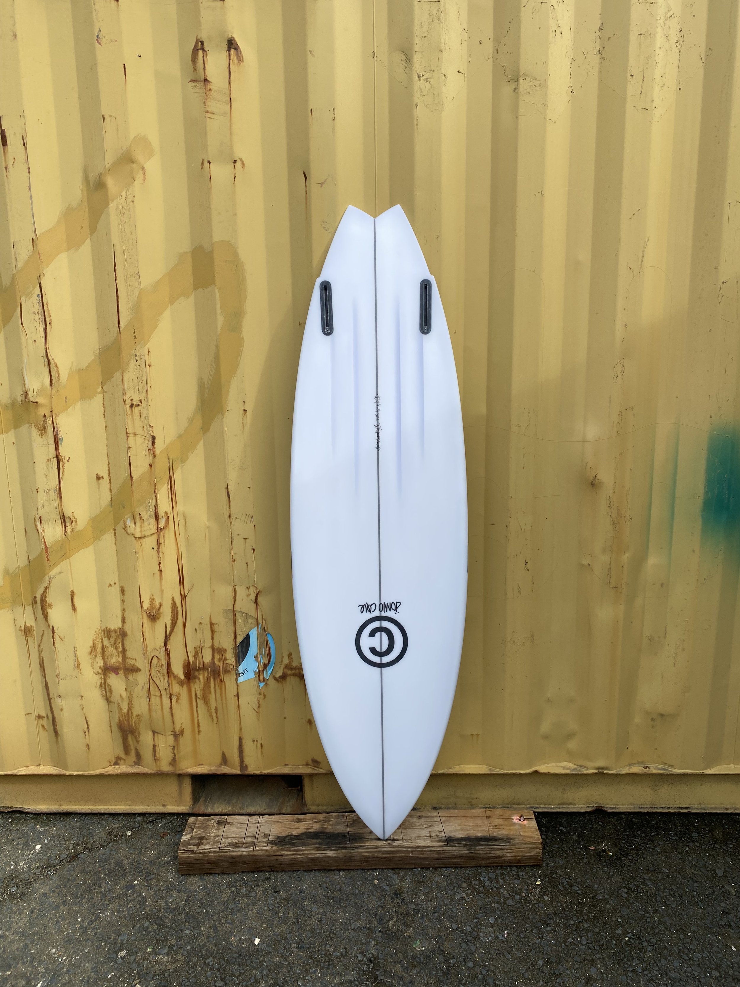 White surfboard with black fins and logo leaning against a yellow rusty shipping container.