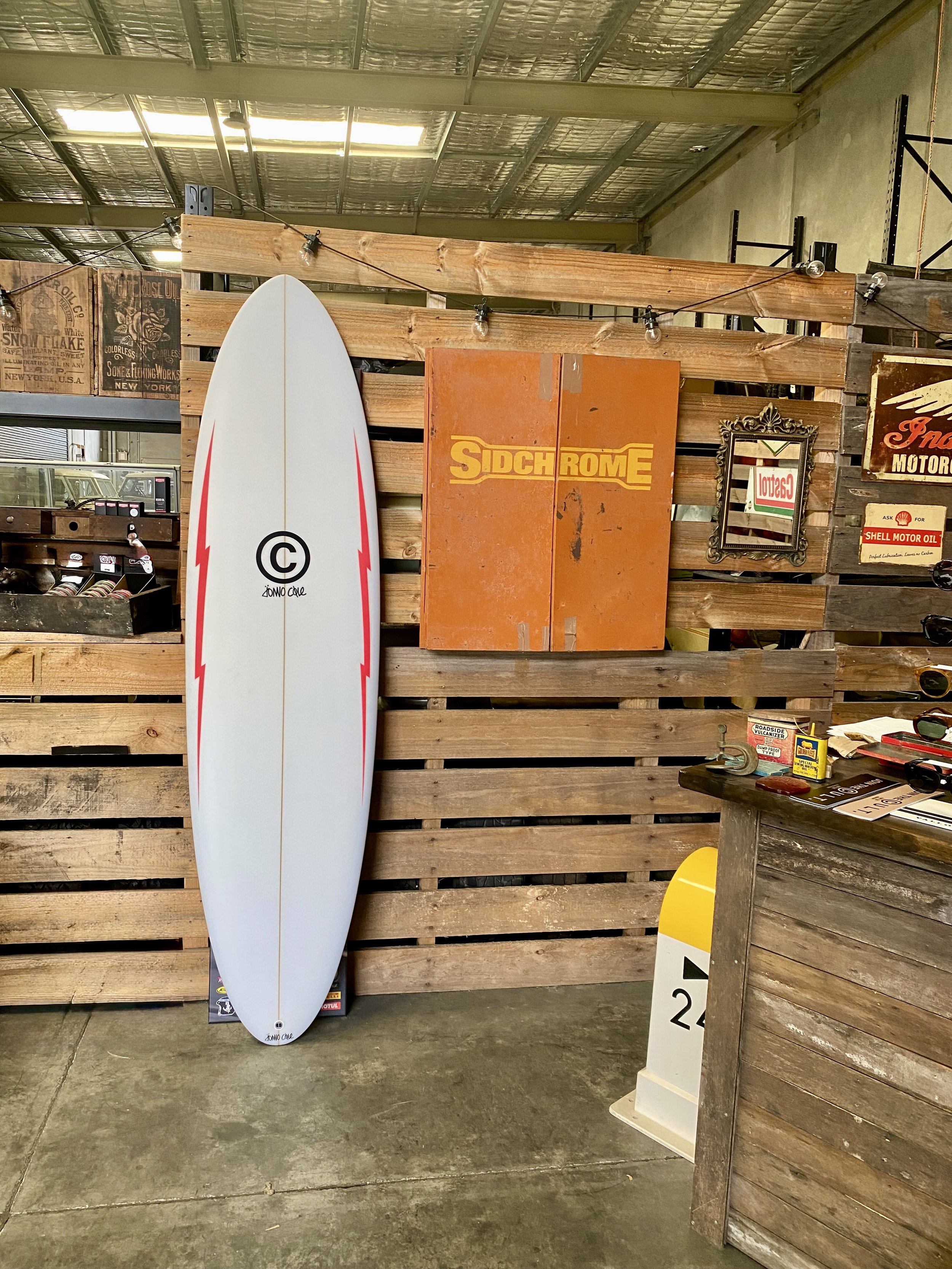 A surfboard with a white background and red lightning bolt designs leaning against a wooden wall. The wall has vintage signs and a mirror, with a rustic wooden counter on the right side holding miscellaneous items. A yellow and white road reflector is on the floor next to the counter.