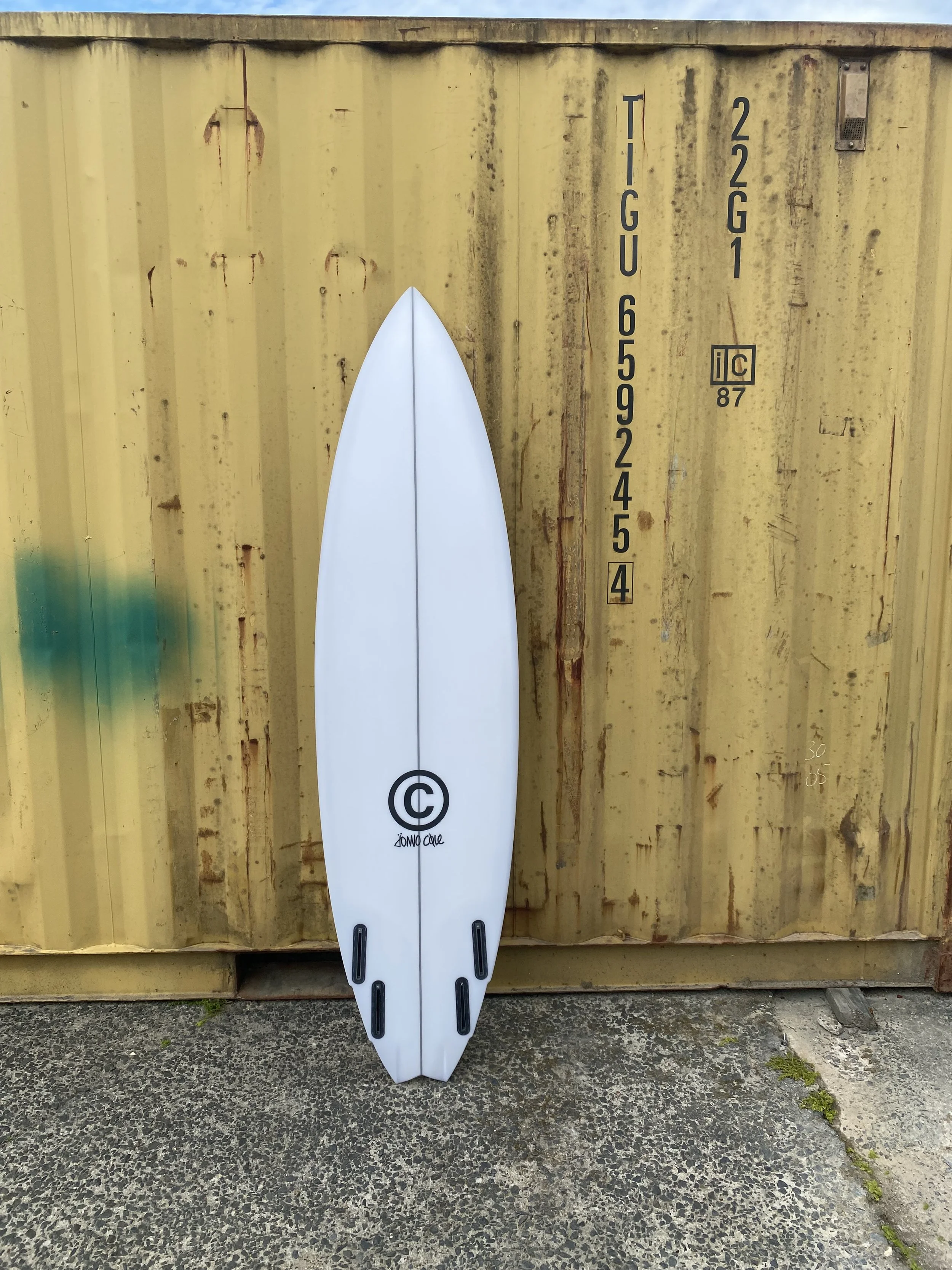 White surfboard with black traction pads leaning against a yellow shipping container.