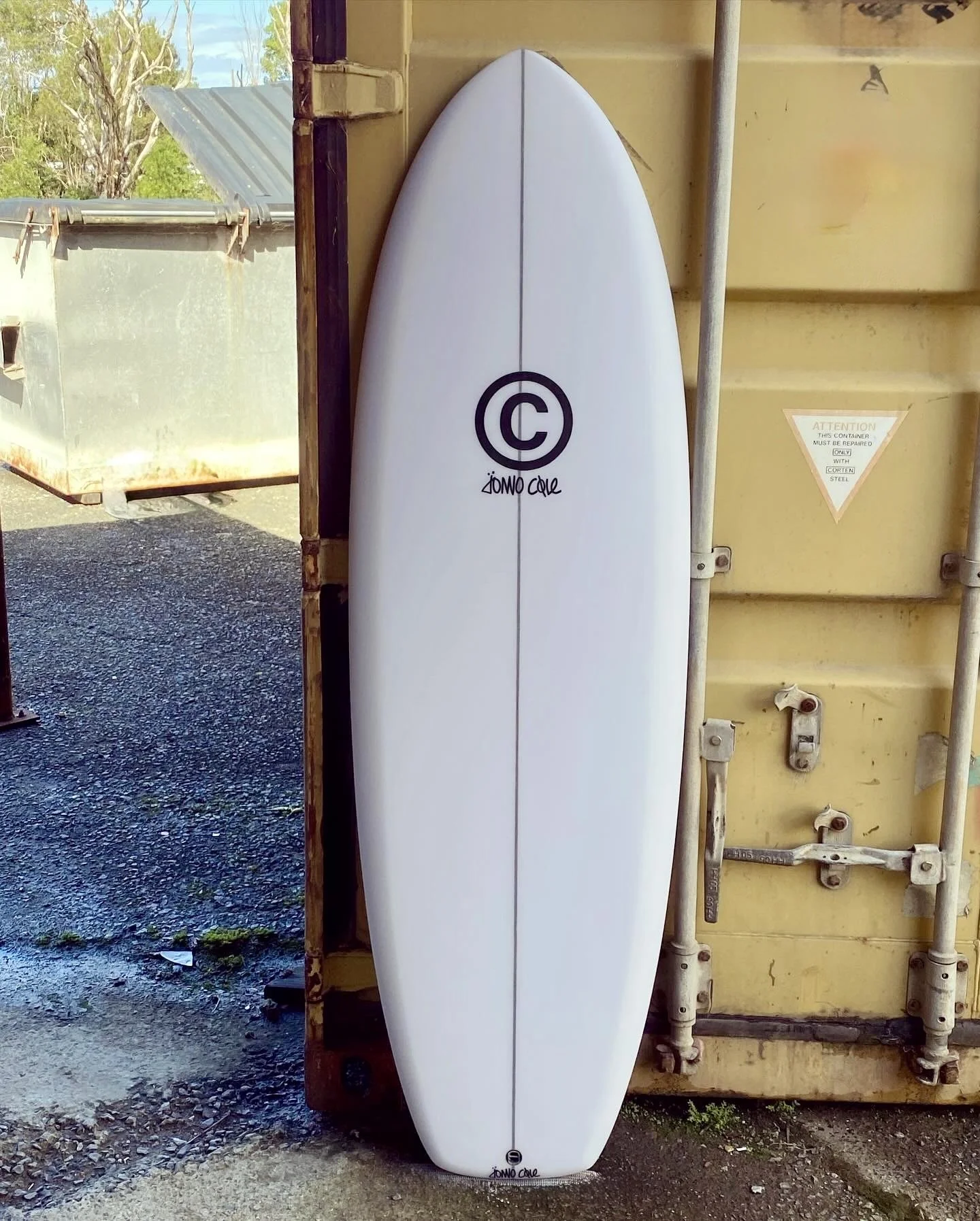 A white surfboard leaning against a yellow shipping container, with a black logo and signature on it.