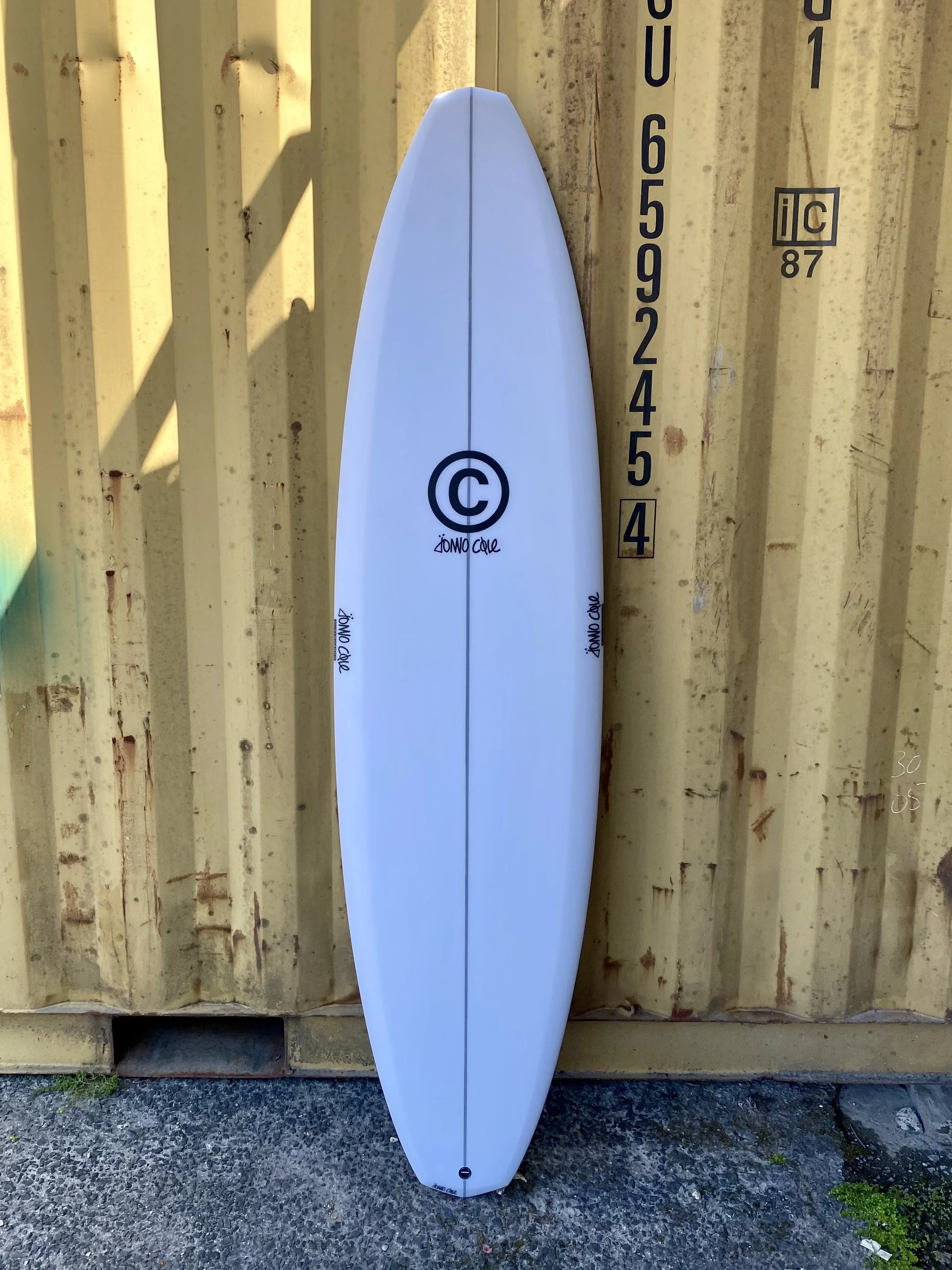 A white surfboard standing upright against a yellow shipping container with black measurement markings.
