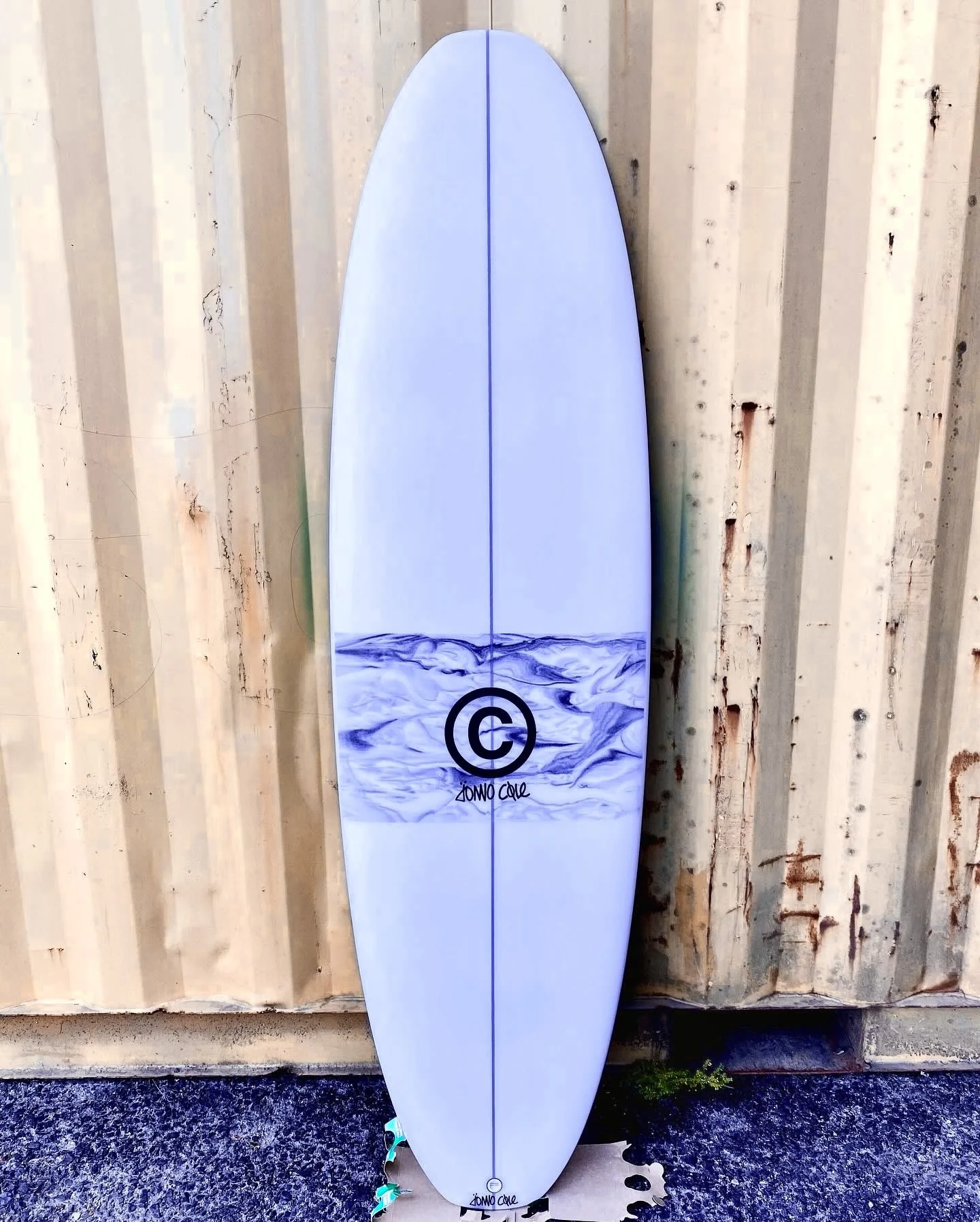 A white surfboard with a blue water wave design and a black copyright symbol, leaning against a wooden wall.