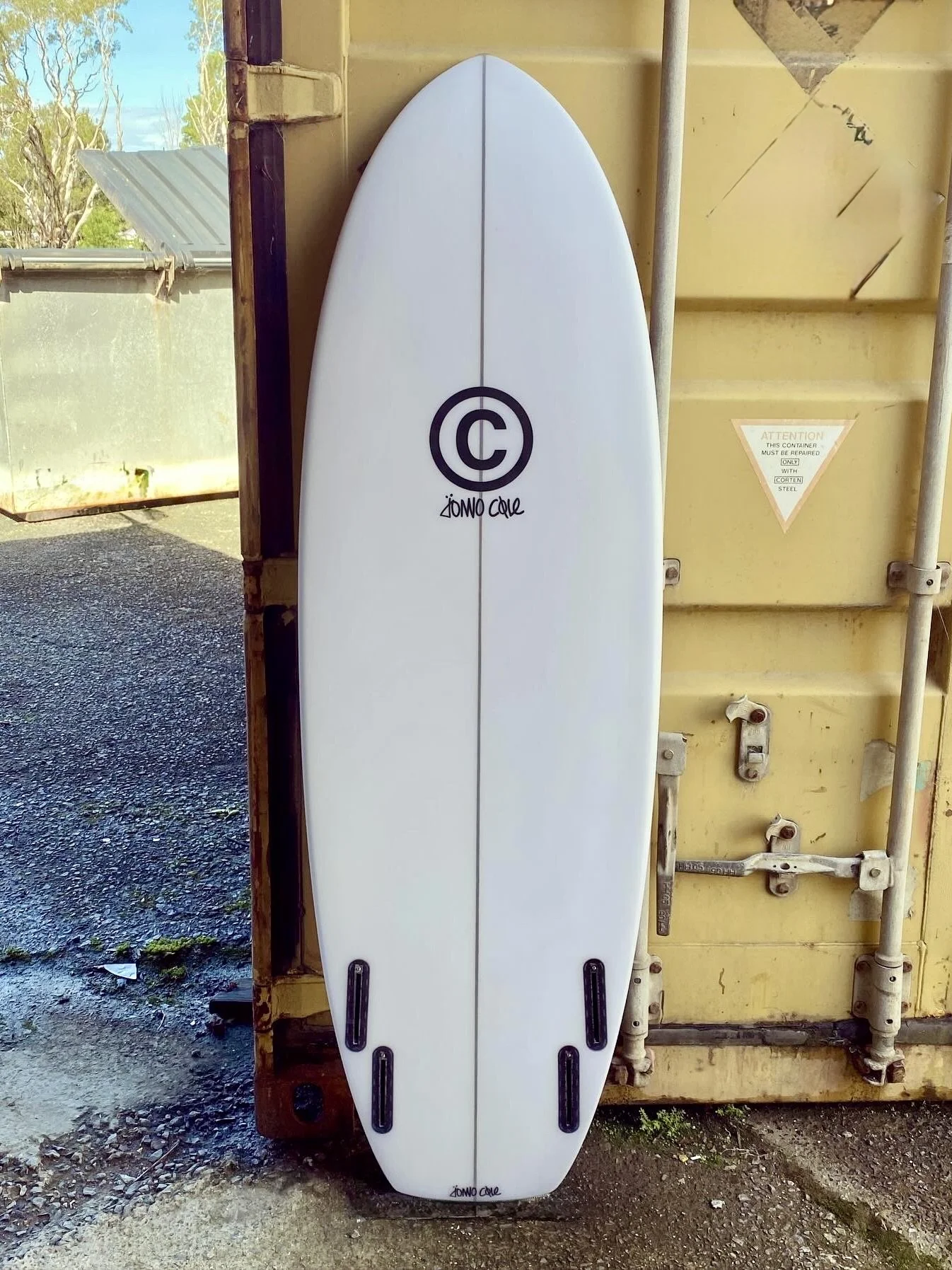 White surfboard with a black circular logo and the words 'Zomo Cole' printed on it, leaning against a yellow shipping container.