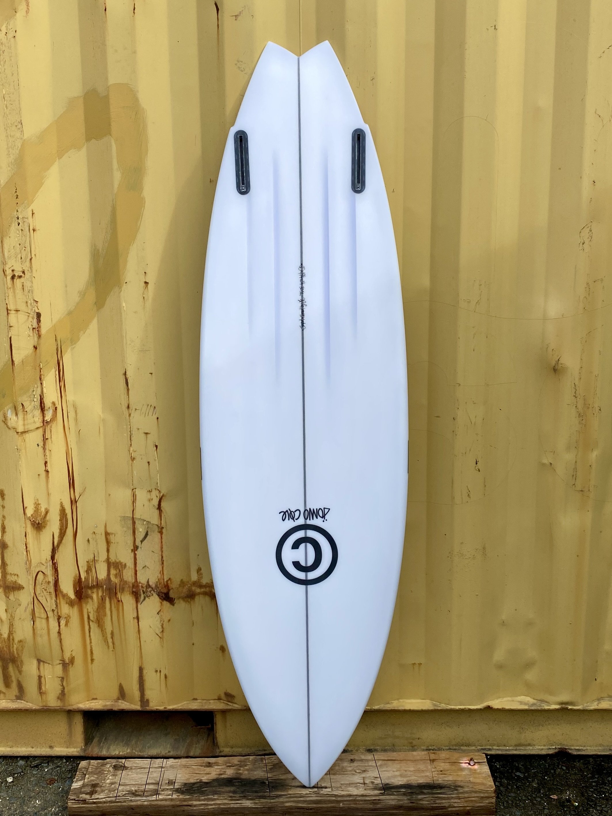 A white surfboard with black fin attachments leaning against a yellow wooden wall.