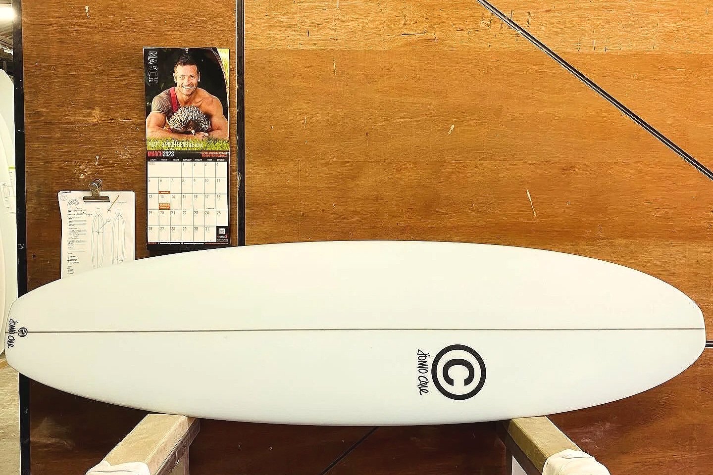 A white foam surfboard with a black logo and copyright symbol, placed on a wooden table with a calendar and other papers in the background.