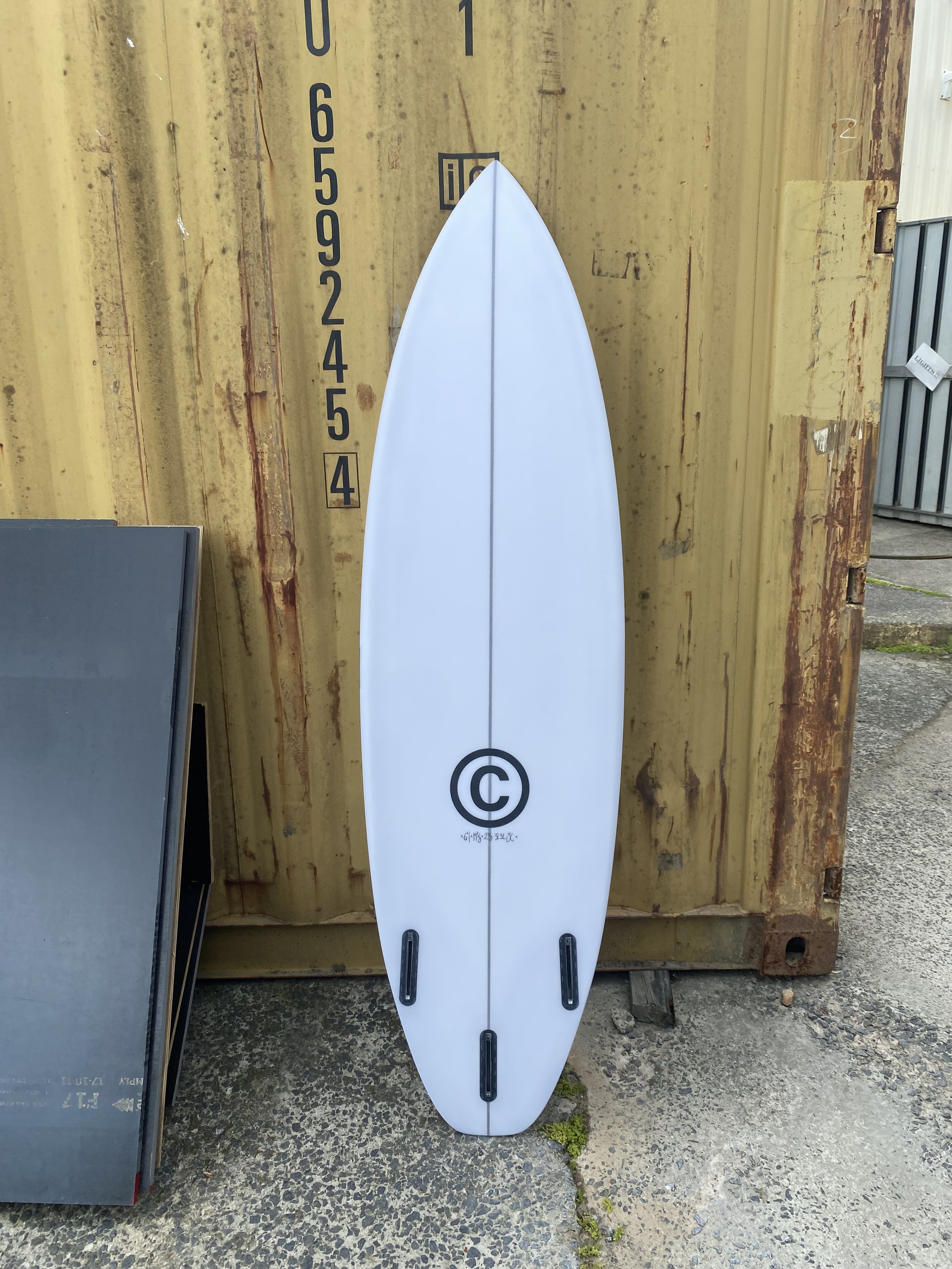 White surfboard with black traction pads and a copyright symbol logo, standing upright against a yellow shipping container.