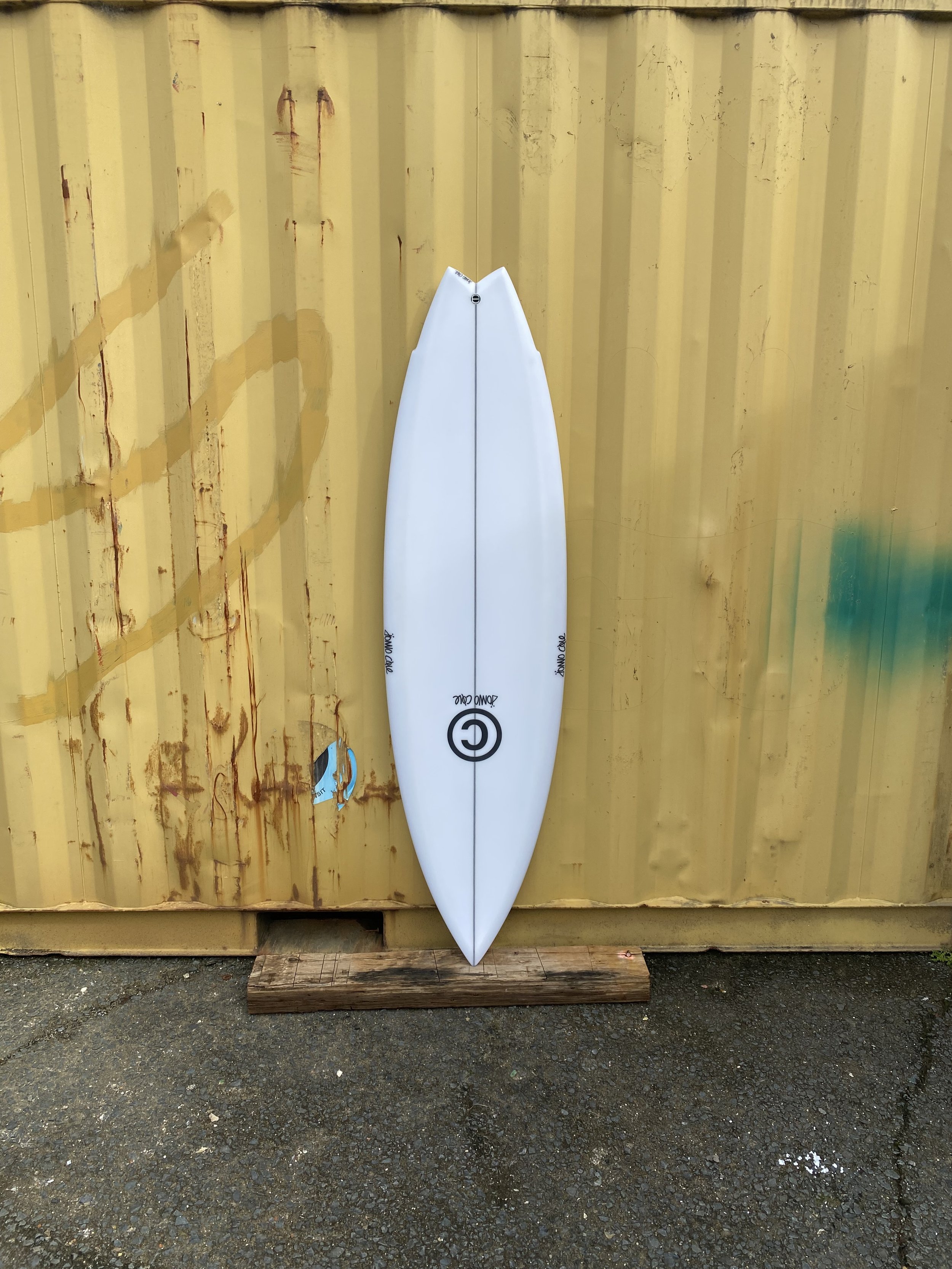 A white surfboard with black logo and text, leaning against a yellow corrugated metal wall, mounted on a small wooden plank on wet pavement.