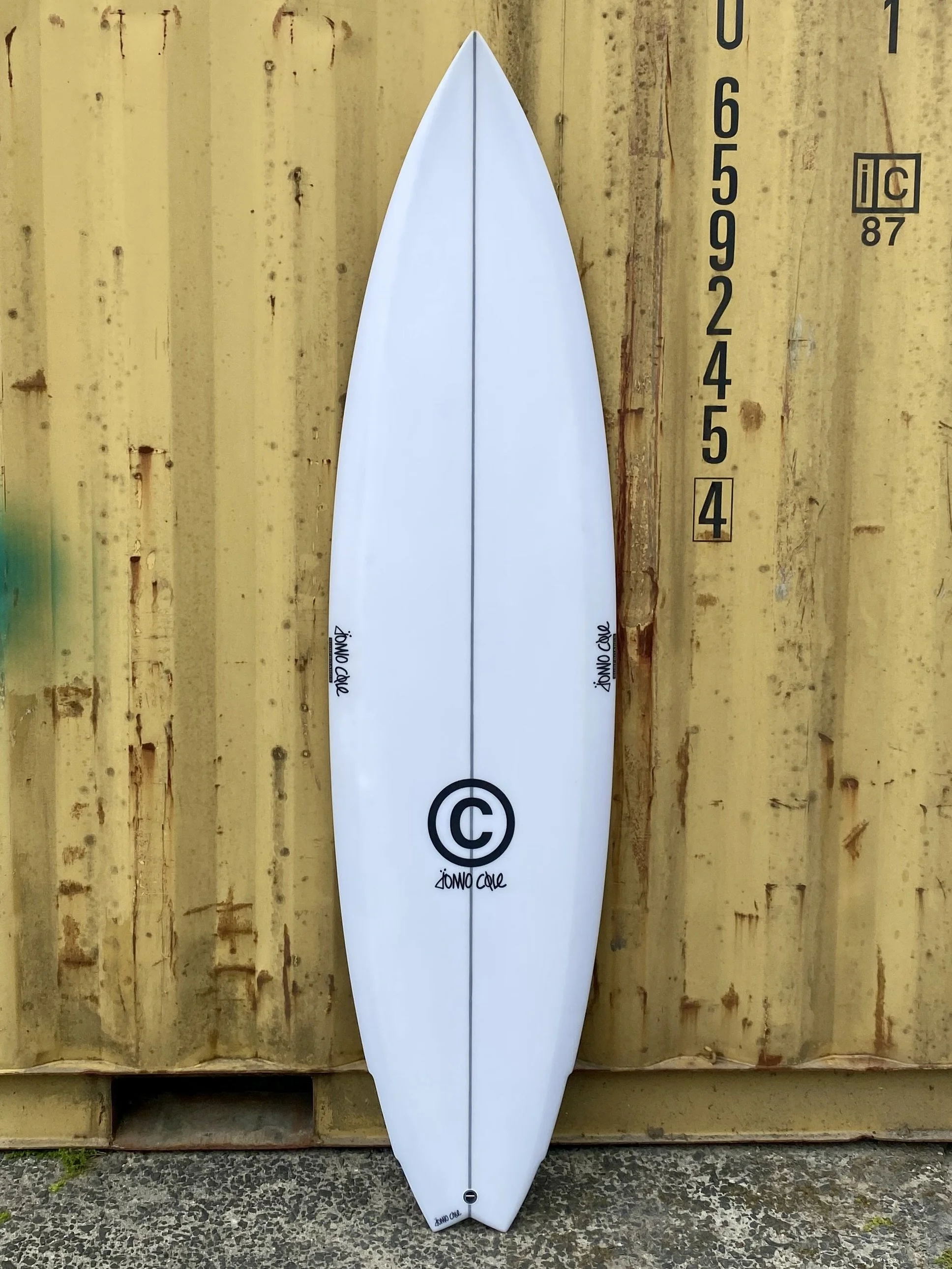 White surfboard with black and white logo leaning against a yellow corrugated metal wall.