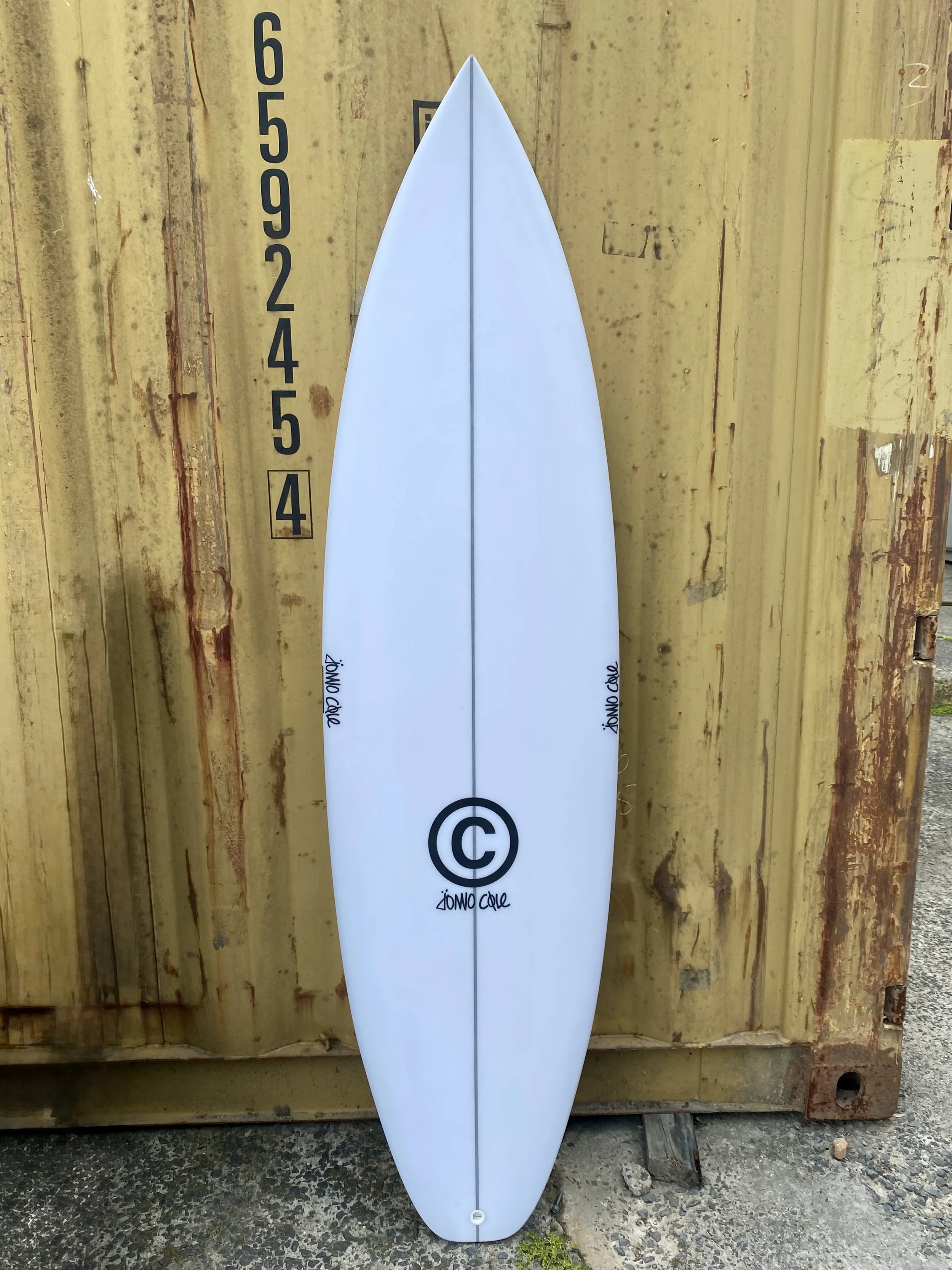 White surfboard with black logo and signature leaning against a wooden wall with height markings.