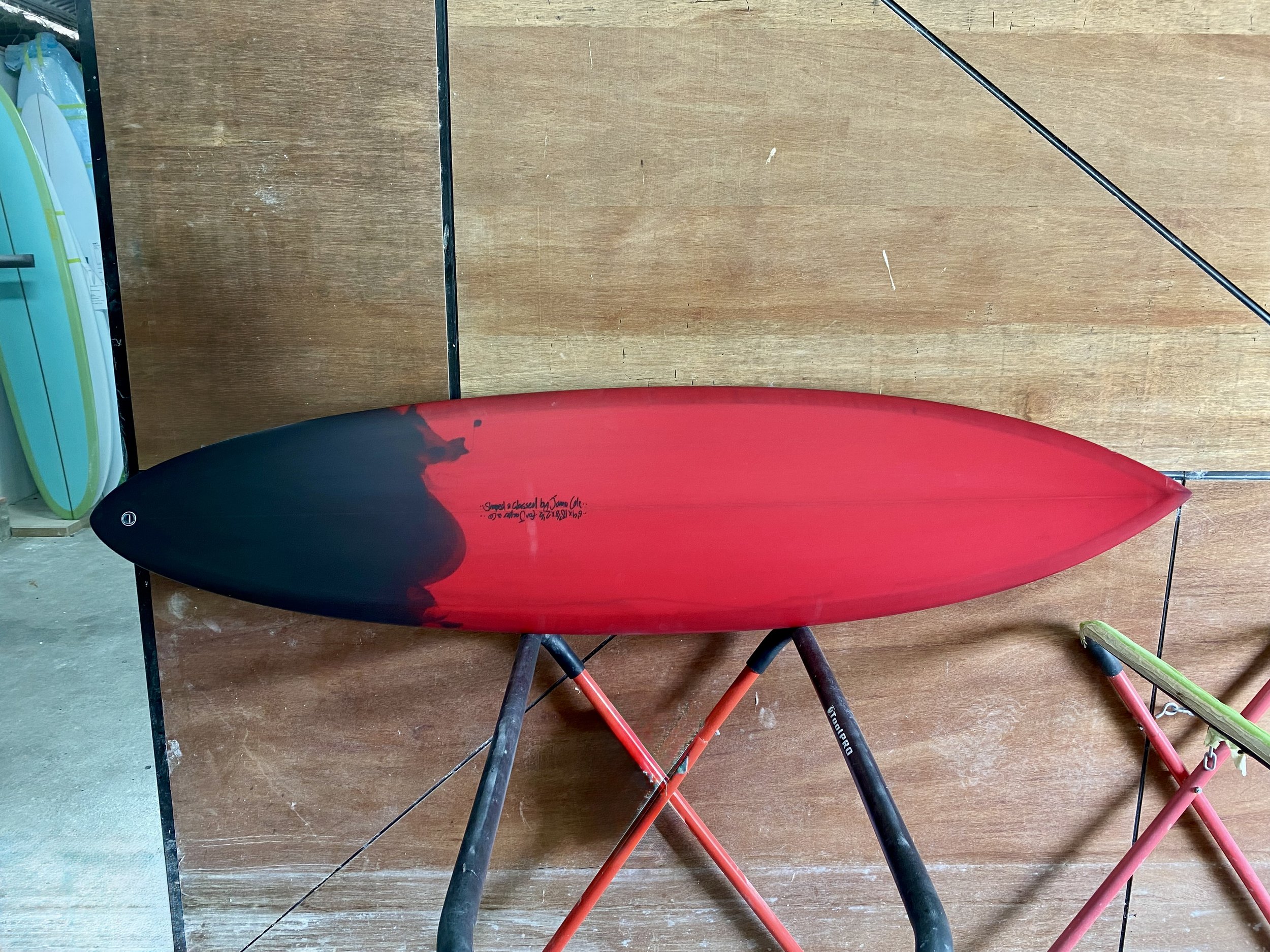 A red and black surfboard resting on a black metal stand against a wood-paneled wall.