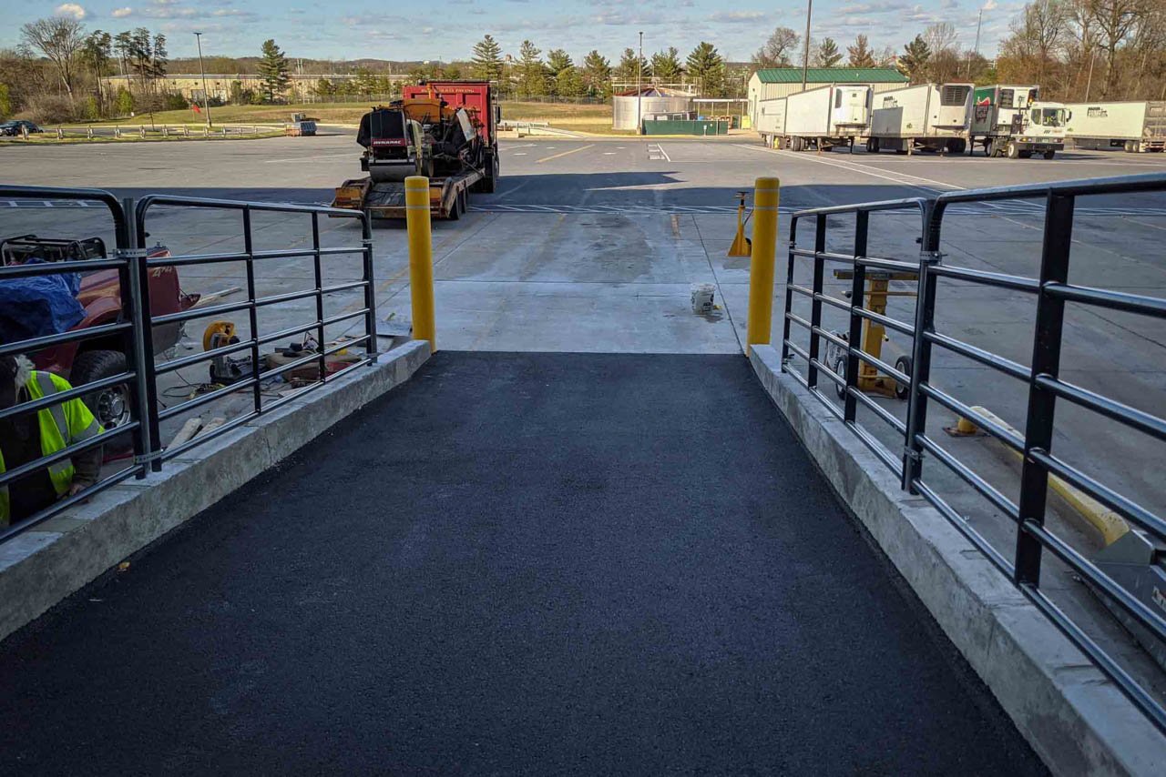 View from a ramp leading to a loading dock area with trucks, trailers, and industrial equipment. Railings and yellow safety bollards are present.