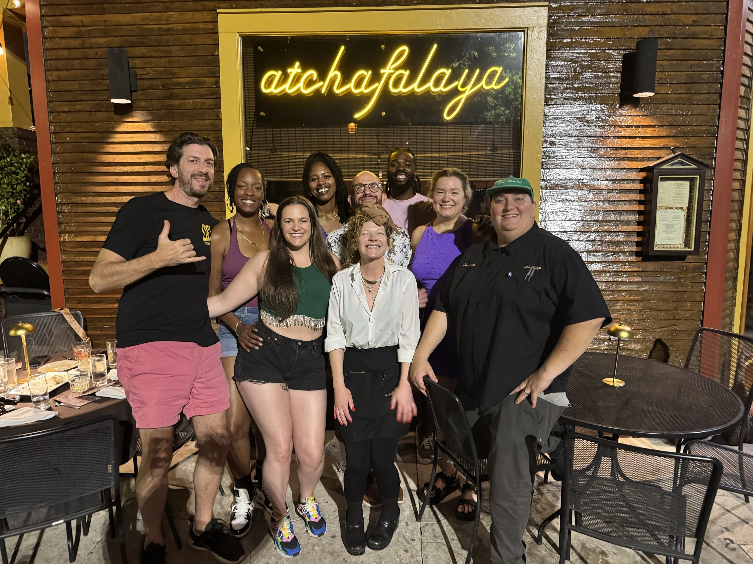 A group of 9 people — male female black white old young — gather in front of a restaurant with happy full smiles. Six are the birthday party; the chef, server, and general manager join the shot.