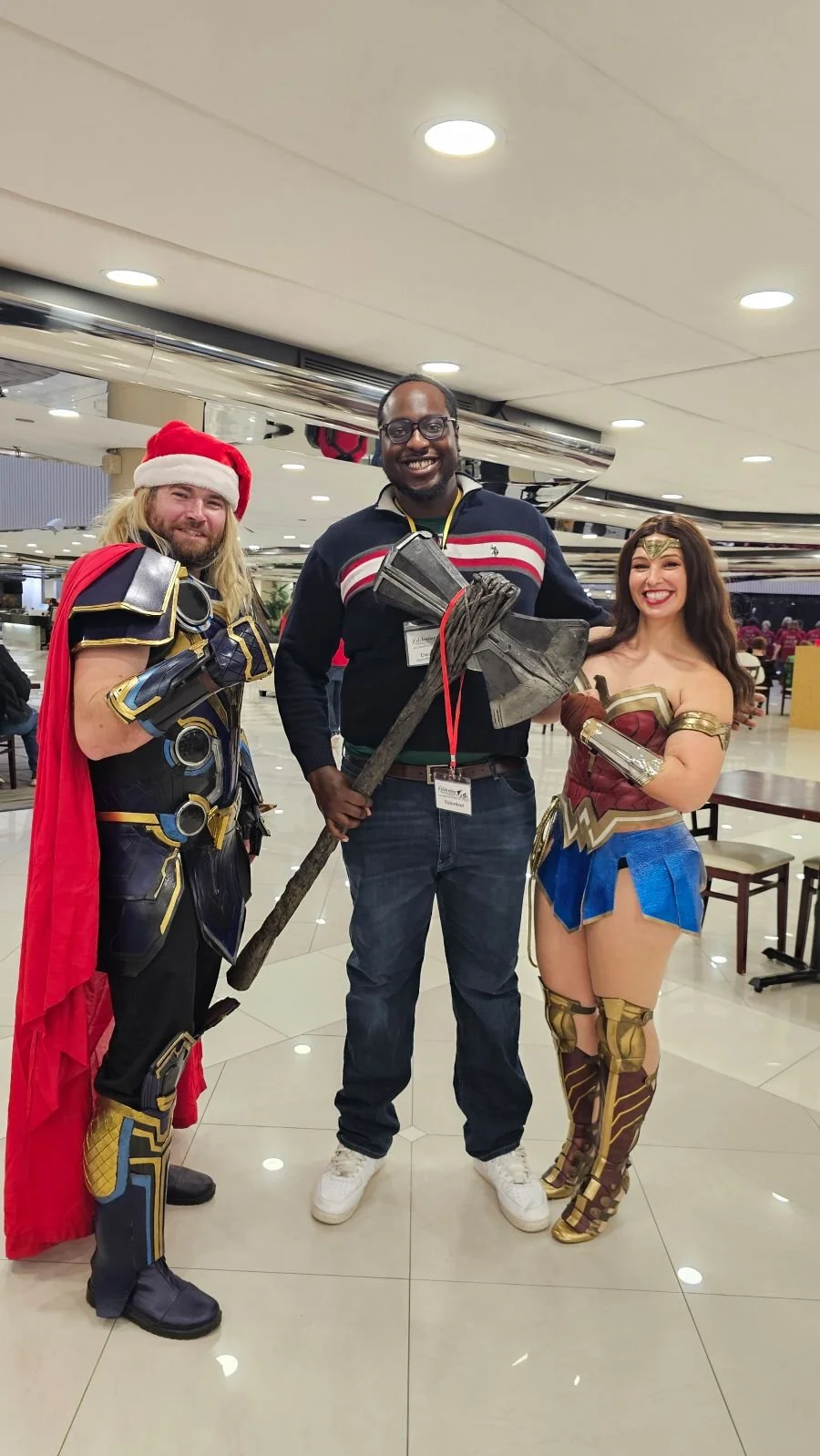 Three people in cosplay costumes posing together indoors; one dressed as Thor with a red cape and Santa hat, the middle person with a Viking axe, and the third as Wonder Woman with a red, blue, and gold costume.