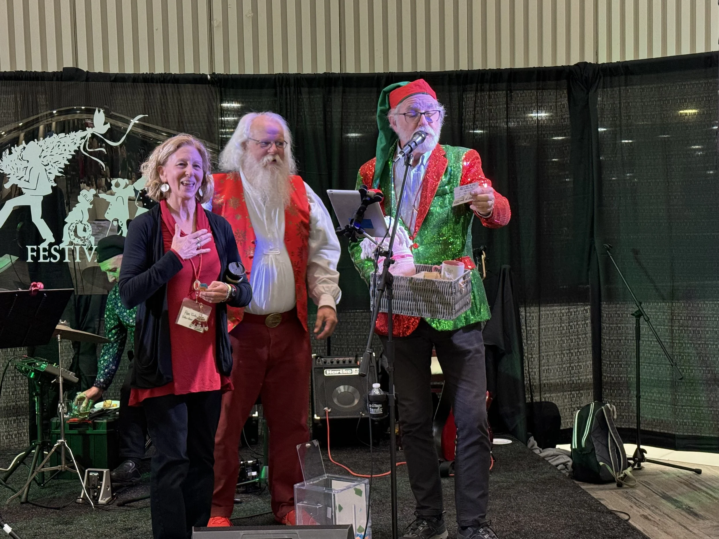 Three people dressed in Christmas-themed attire on a stage with musical equipment, one speaking into a microphone, during a festive event.