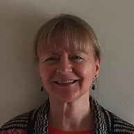 A smiling woman with short blonde hair, wearing a red top and a patterned cardigan, standing indoors.