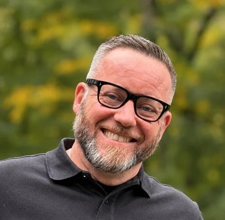 A man with glasses and a beard smiling, wearing a dark gray sweater over a maroon collared shirt, standing against a plain gray background.