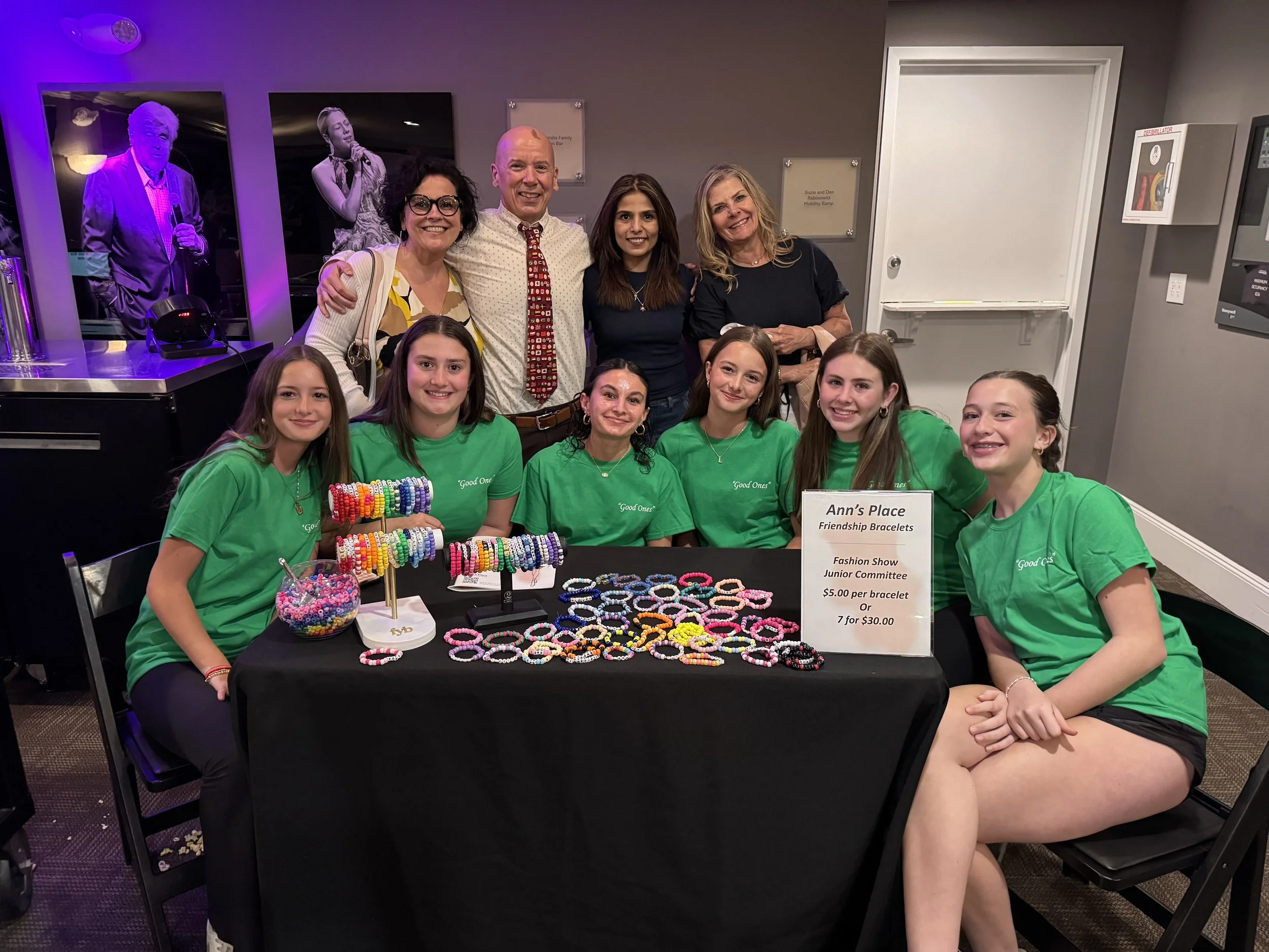 A group of young girls in green shirts sitting at a table selling friendship bracelets, with a sign indicating prices, and a few adults standing behind them, in an indoor setting with photographs on the wall.