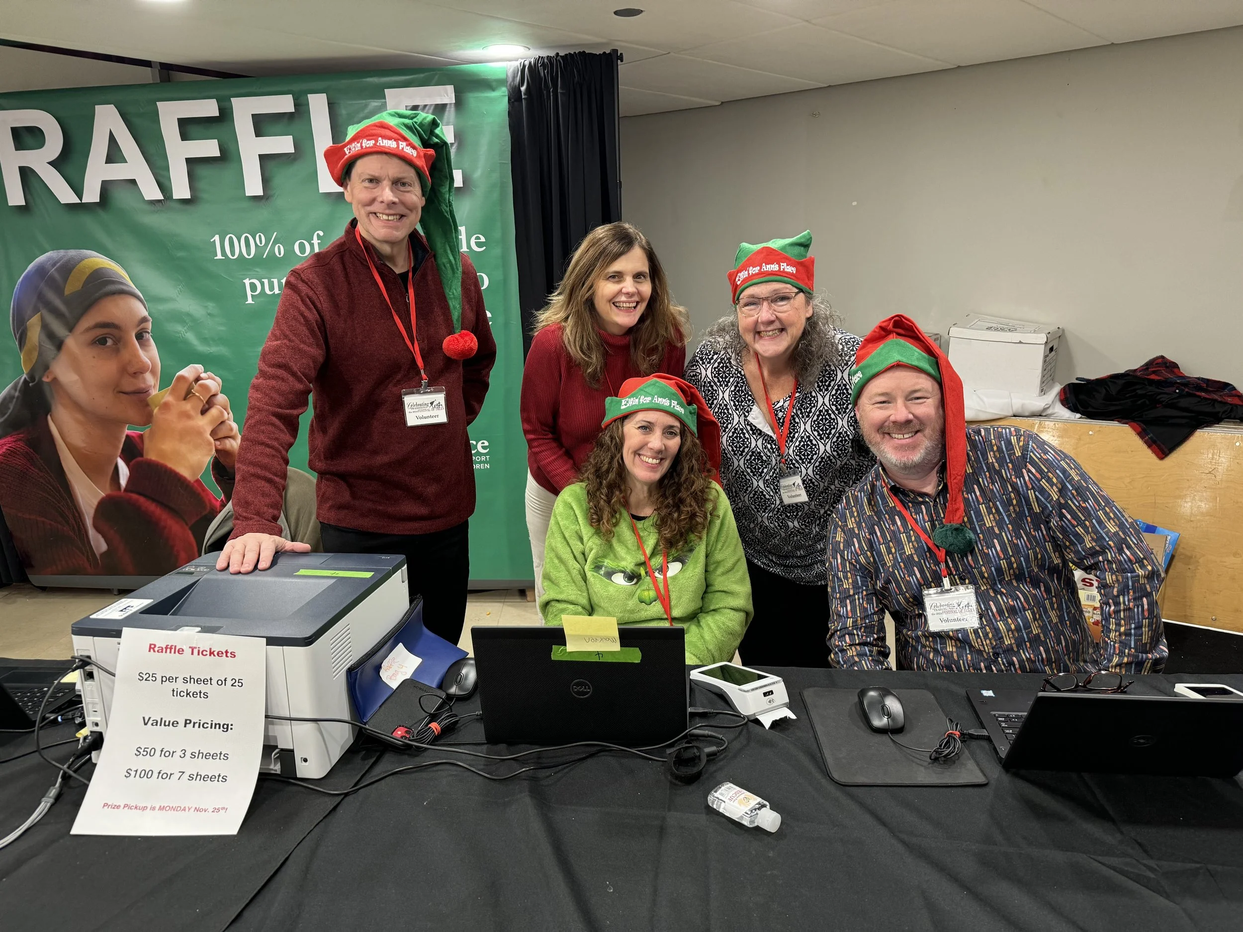Group of six people at a Christmas-themed event, wearing elf hats and holiday attire, seated and standing behind a table with raffle tickets and equipment, smiling for the camera.
