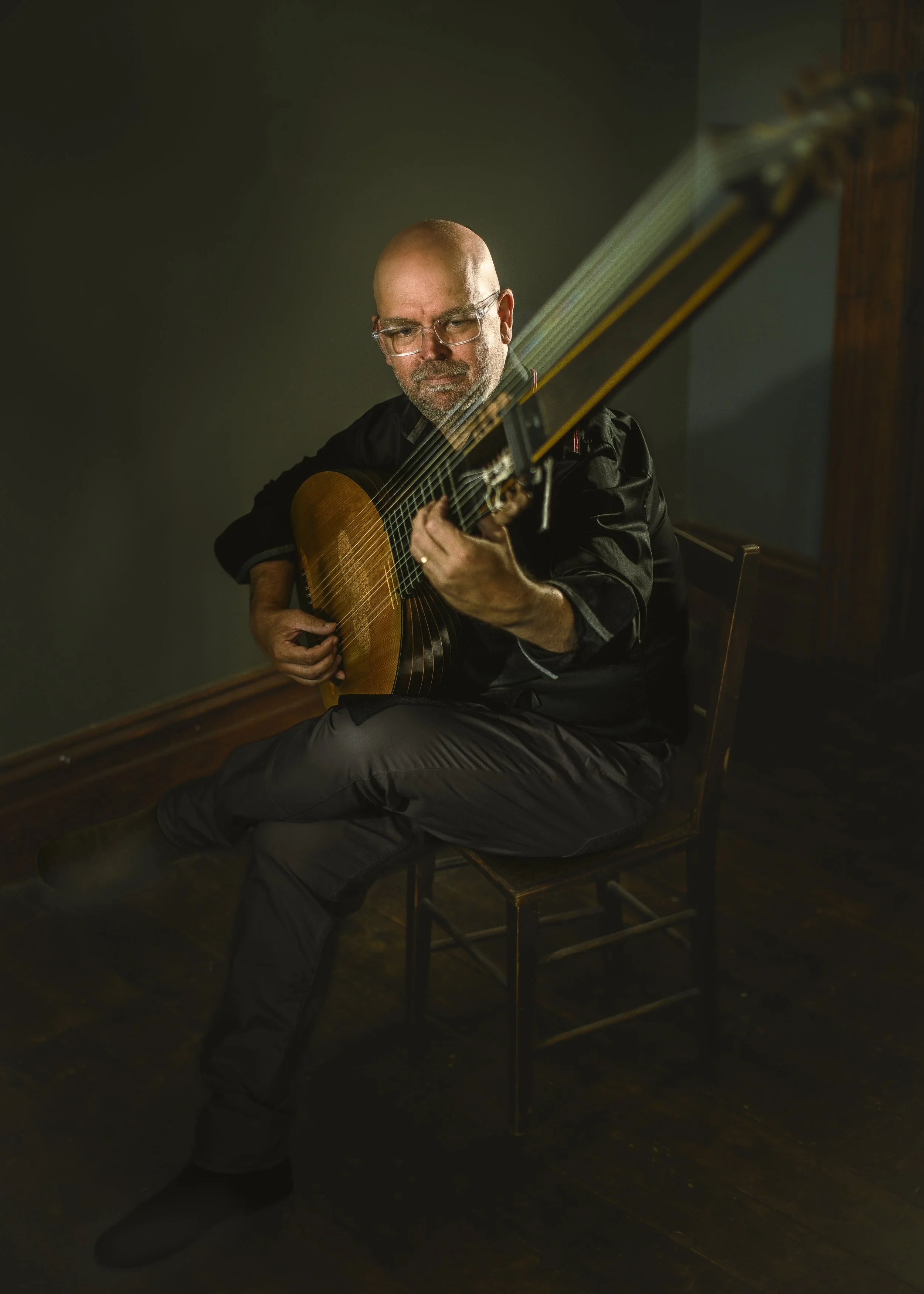 Simon Martyn-Ellis sitting on a chair playing his theorbo. The entire length of the theorbo neck is in view, but out of focus.