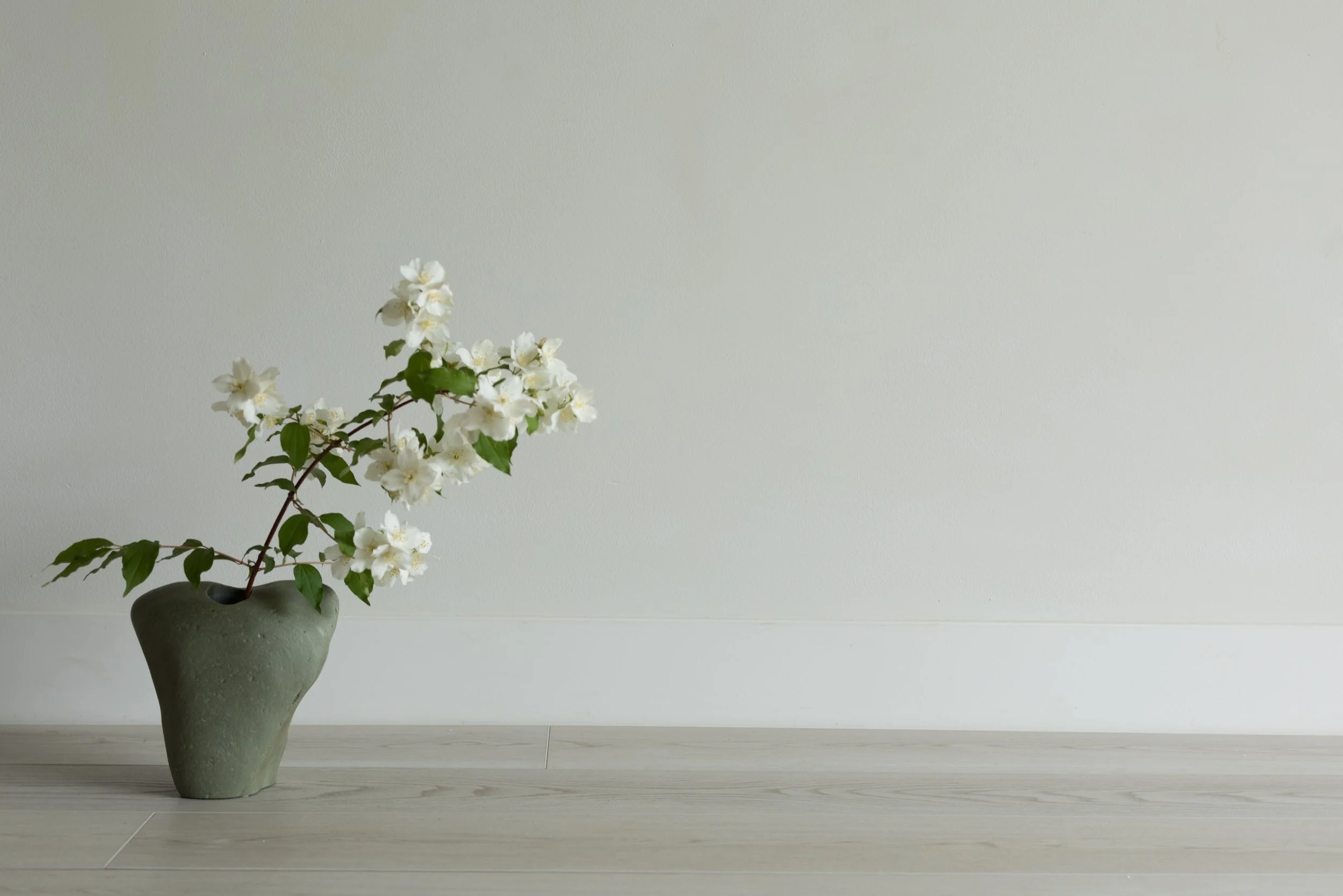 White flowers in a green ceramic vase on a wooden floor against a light wall.