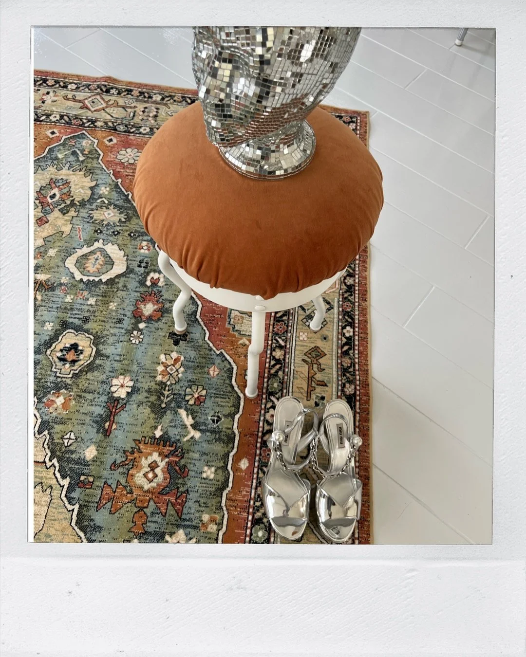 Decorative interior scene with a patterned rug, a stool with a brown cushion, a mirrored bust on the stool, and metallic high-heeled sandals.