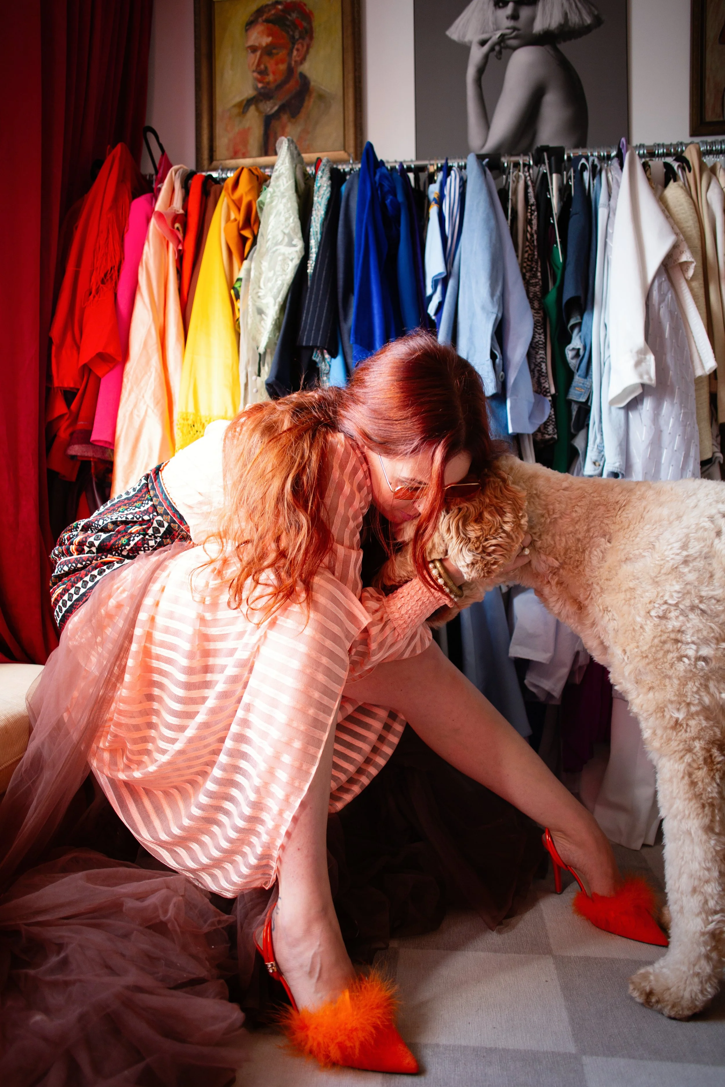 Woman wearing a dress. Sitting in a closet with bright clothing. She is hugging her golden doodle dog.