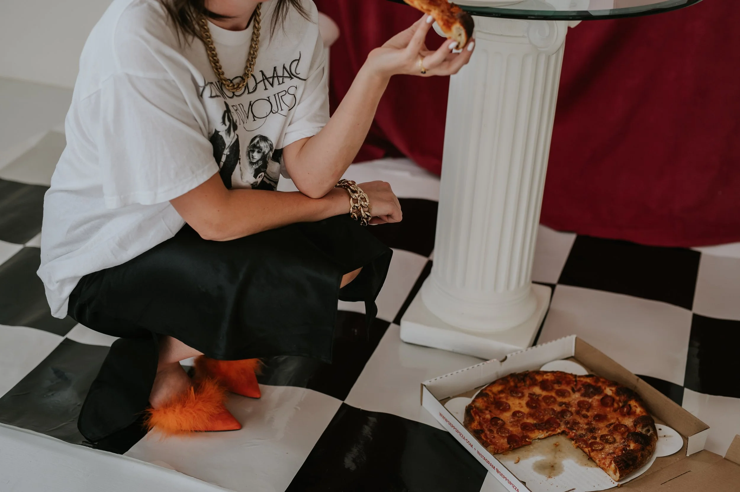 Person in a white graphic t-shirt and orange feathered shoes, holding a slice of pepperoni pizza beside a pizza box on a black and white checkered floor, next to a white pedestal table.