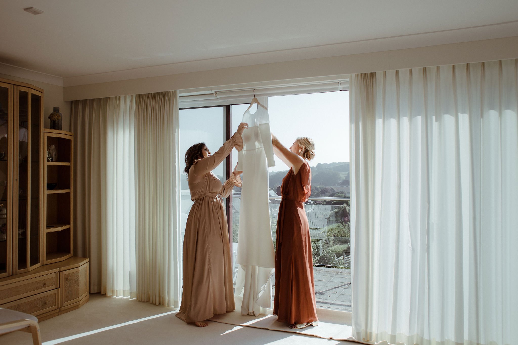 Bridesmaids, dressed in terracotta bridesmaid dresses, in window taking wedding dress off hanger