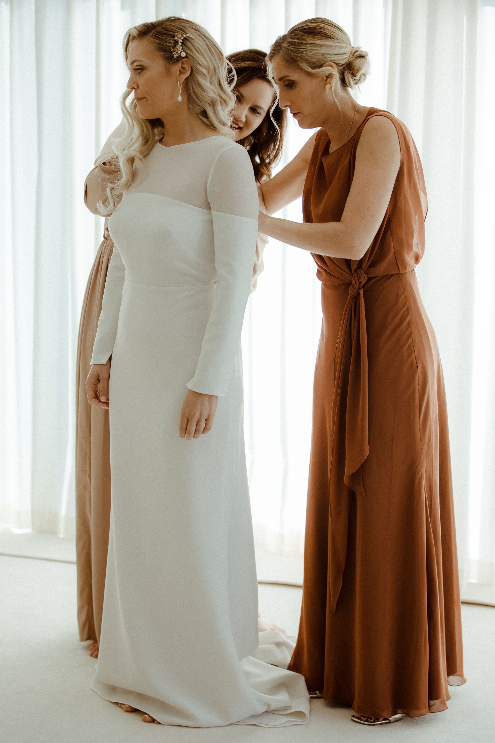 Bridesmaids dressed in terracotta dresses helping contemporary bride into her wedding dress with sleeves for October wedding