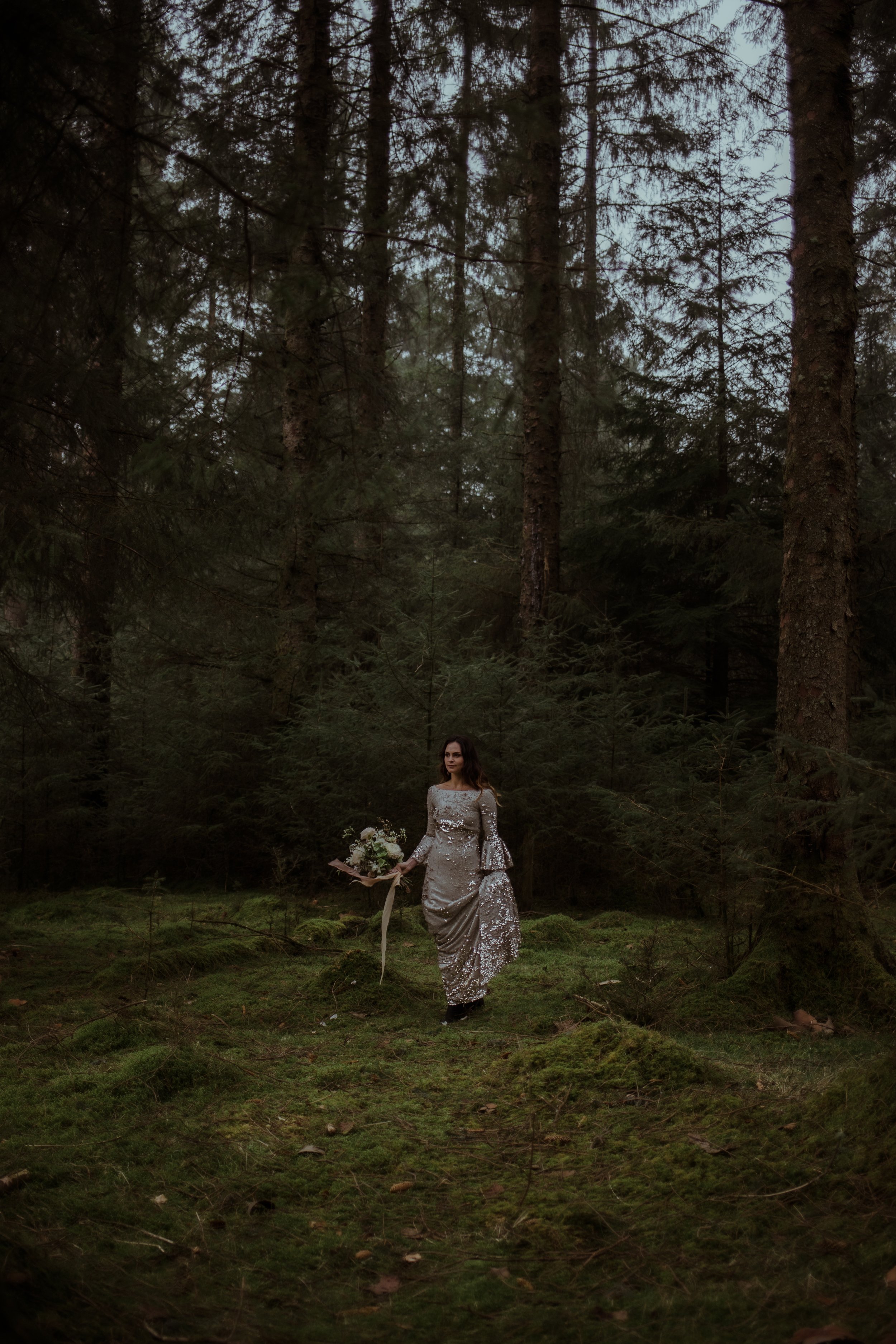 Bride standing in dark green forest wearing silver sequin wedding dress