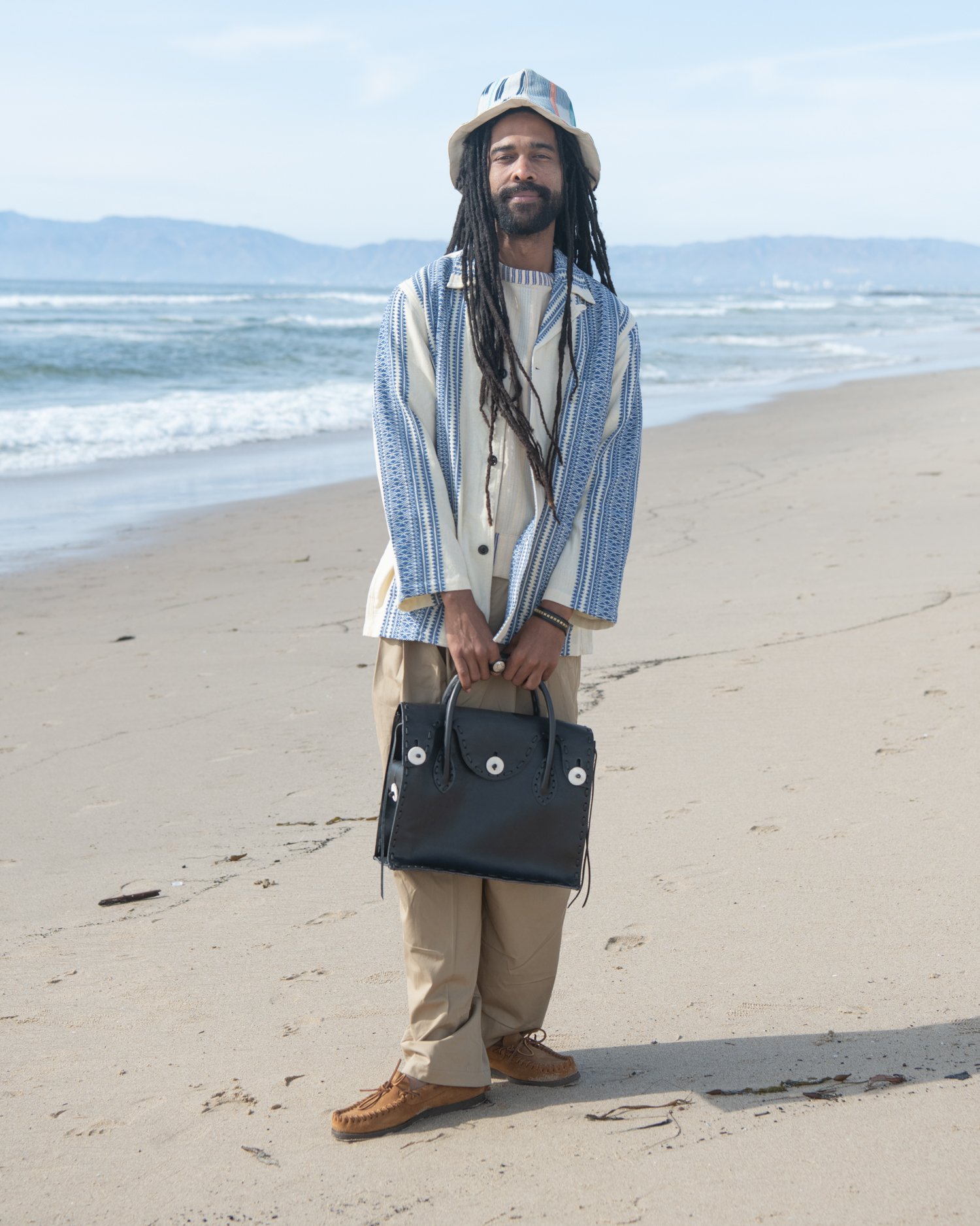 The Italian jail jacket. Featuring an indigo and natural hand-loomed cotton jacquard being worn by a man on the beach holding Chamula handbag