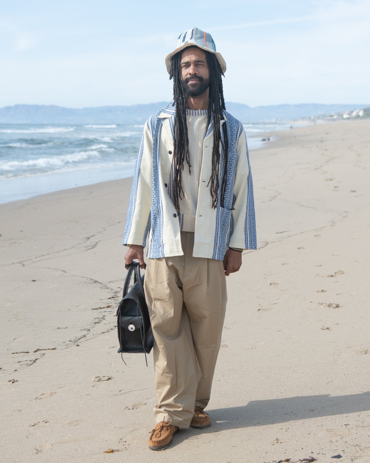The Italian jail jacket. Featuring an indigo and natural hand-loomed cotton jacquard being worn by a man on the beach holding Chamula handbag