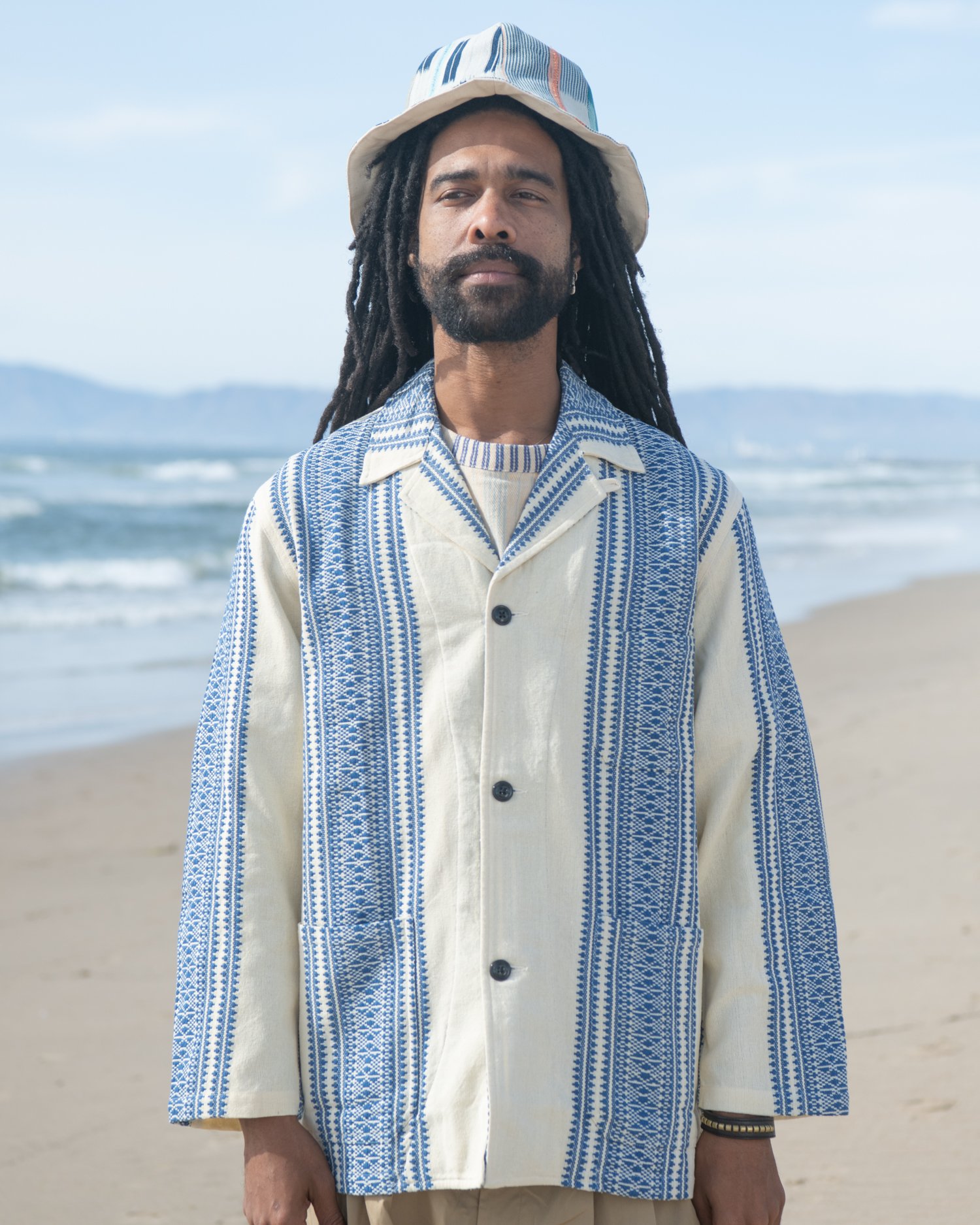 The Italian jail jacket. Featuring an indigo and natural hand-loomed cotton jacquard being worn by a man on the beach holding Chamula handbag