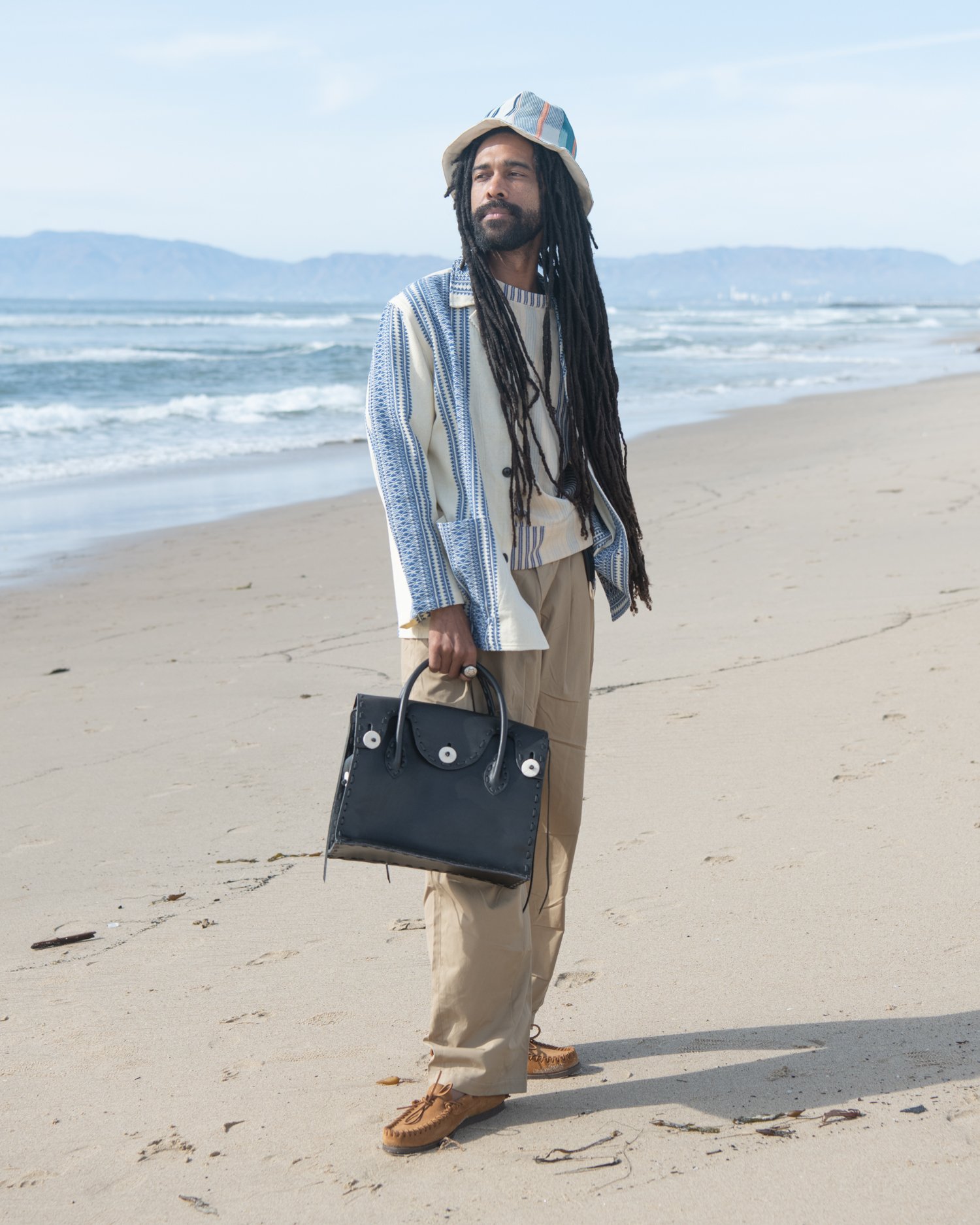 The Italian jail jacket. Featuring an indigo and natural hand-loomed cotton jacquard being worn by a man on the beach holding Chamula handbag