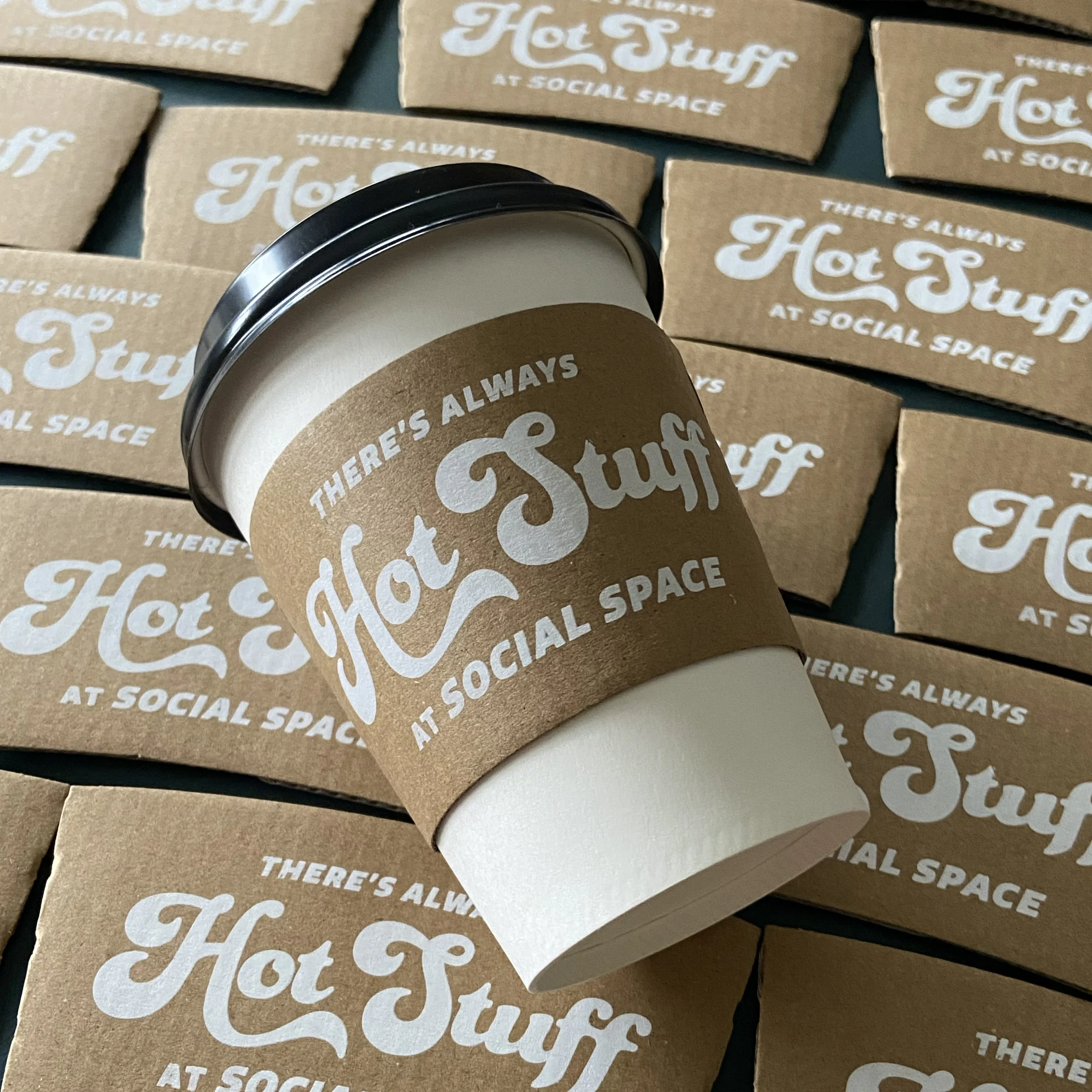 A coffee cup with a brown paper sleeve that says 'There's Always Hot Stuff at Social Space,' surrounded by multiple similar coffee sleeves and boxes with the same branding.