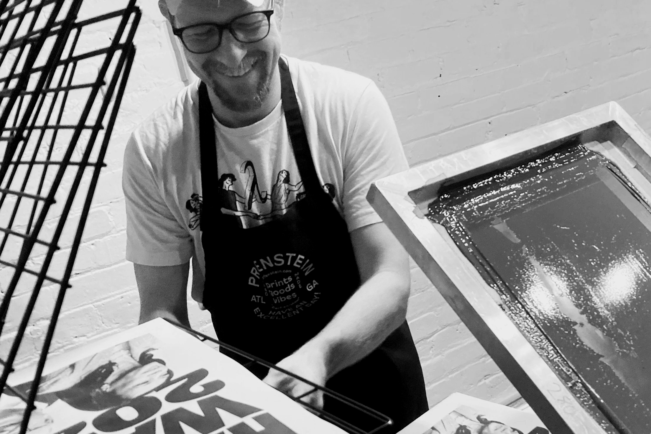 A man with glasses and a beard, smiling, wearing an apron and a t-shirt, working with screen printing equipment in a studio with brick walls.