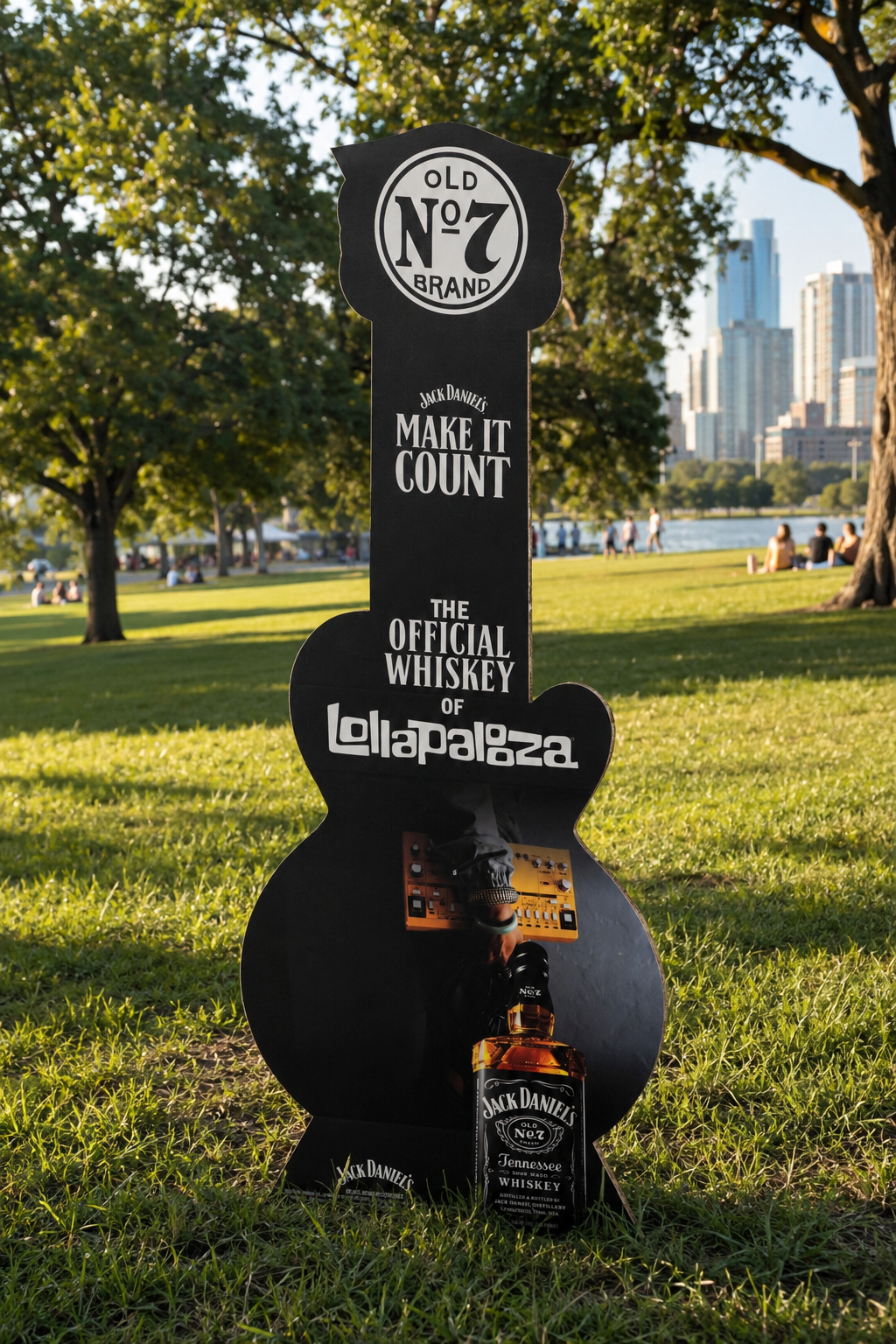A tall, black, guitar-shaped advertisement stand featuring Jack Daniel's whiskey, located outdoors on a grassy field with trees and sunlight in the background.