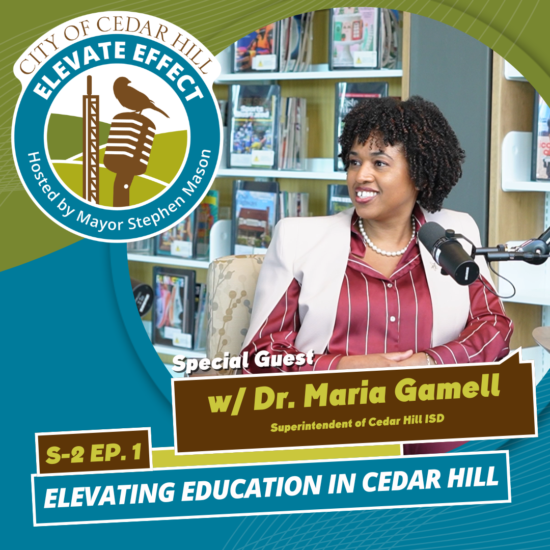 A woman with curly hair sitting at a table in a library or bookstore with books on shelves behind her. She is wearing a white blazer over a red and white striped shirt, with a pearl necklace. She is smiling and speaking into a microphone.