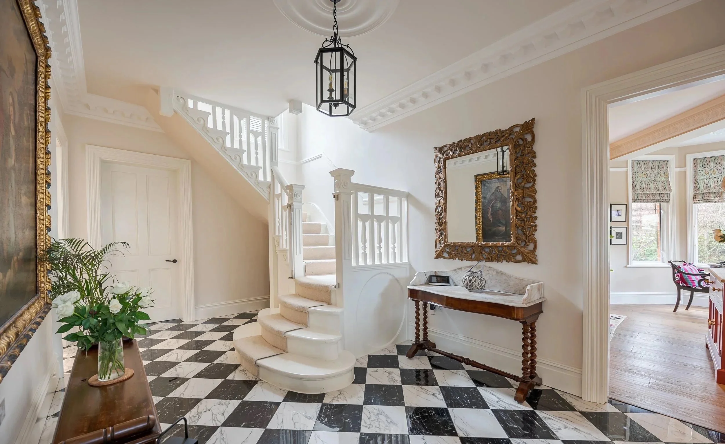 Elegant interior hallway featuring a black and white checkered marble floor, a white staircase with carpeted steps, a wooden console table with decorative items, a large ornate mirror with a gold frame, a houseplant on a small table, and a view into 