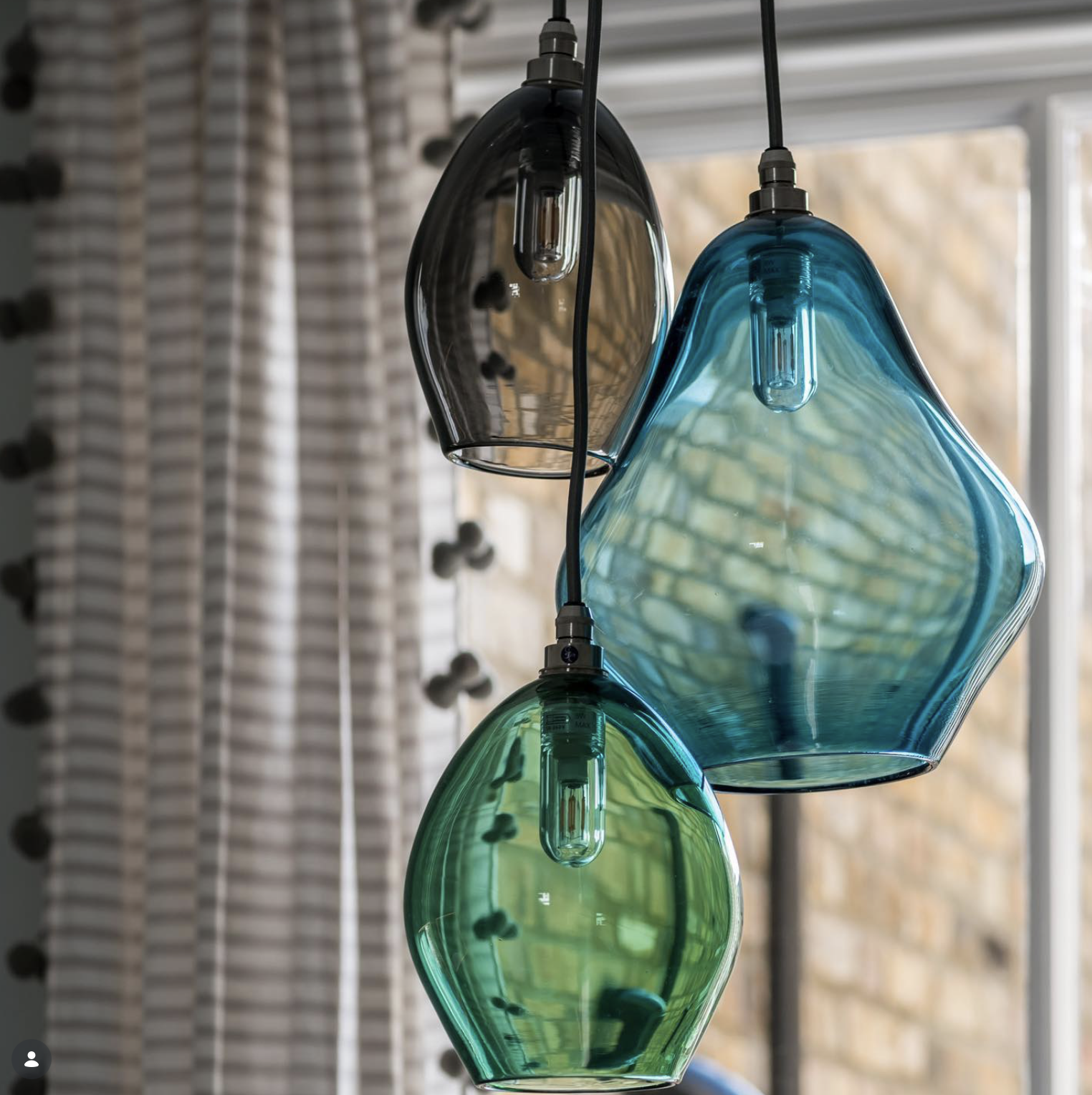 Close-up of three colorful glass pendant lights hanging from the ceiling with a window and curtains in the background.