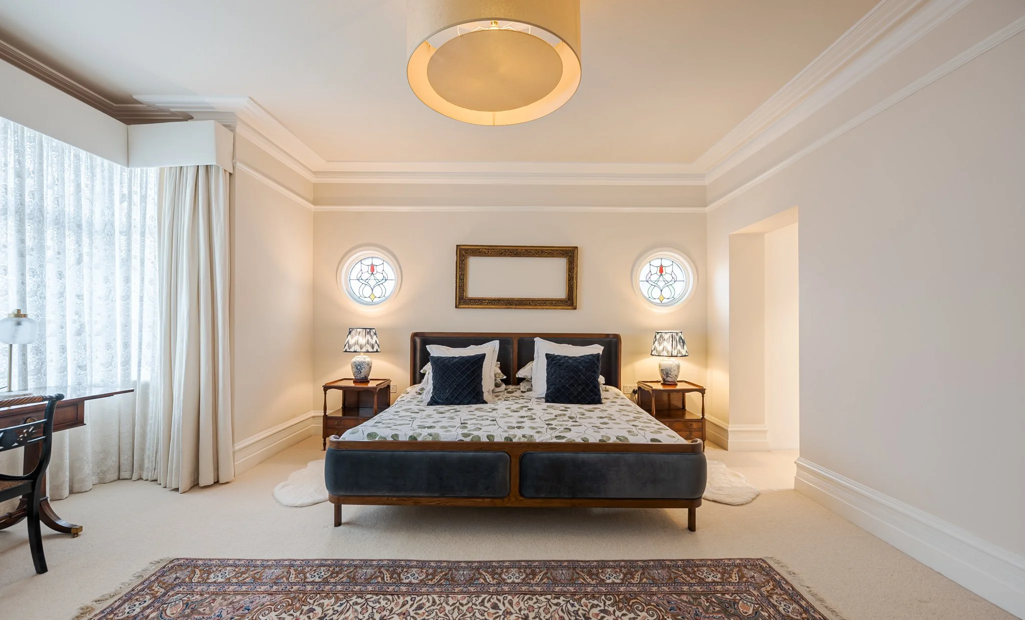 A neatly made bed with a quilted floral bedspread, navy pillows, and white pillows, flanked by two wooden nightstands with lamps, in a well-lit bedroom with stained glass windows, a large ceiling light, a curtain, and an area rug.