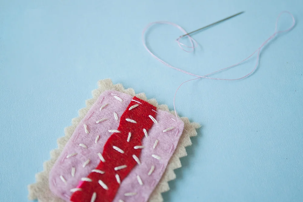 Step 6 To create the biscuit "topping", use three strands of white thread and stitch the red rectangle to the pink rectangle using single stitches, sewing in all directions so it looks like desiccated coconut. Sew the topping to the cream rectangle along the edges using running stitch and a single strand of pink thread.