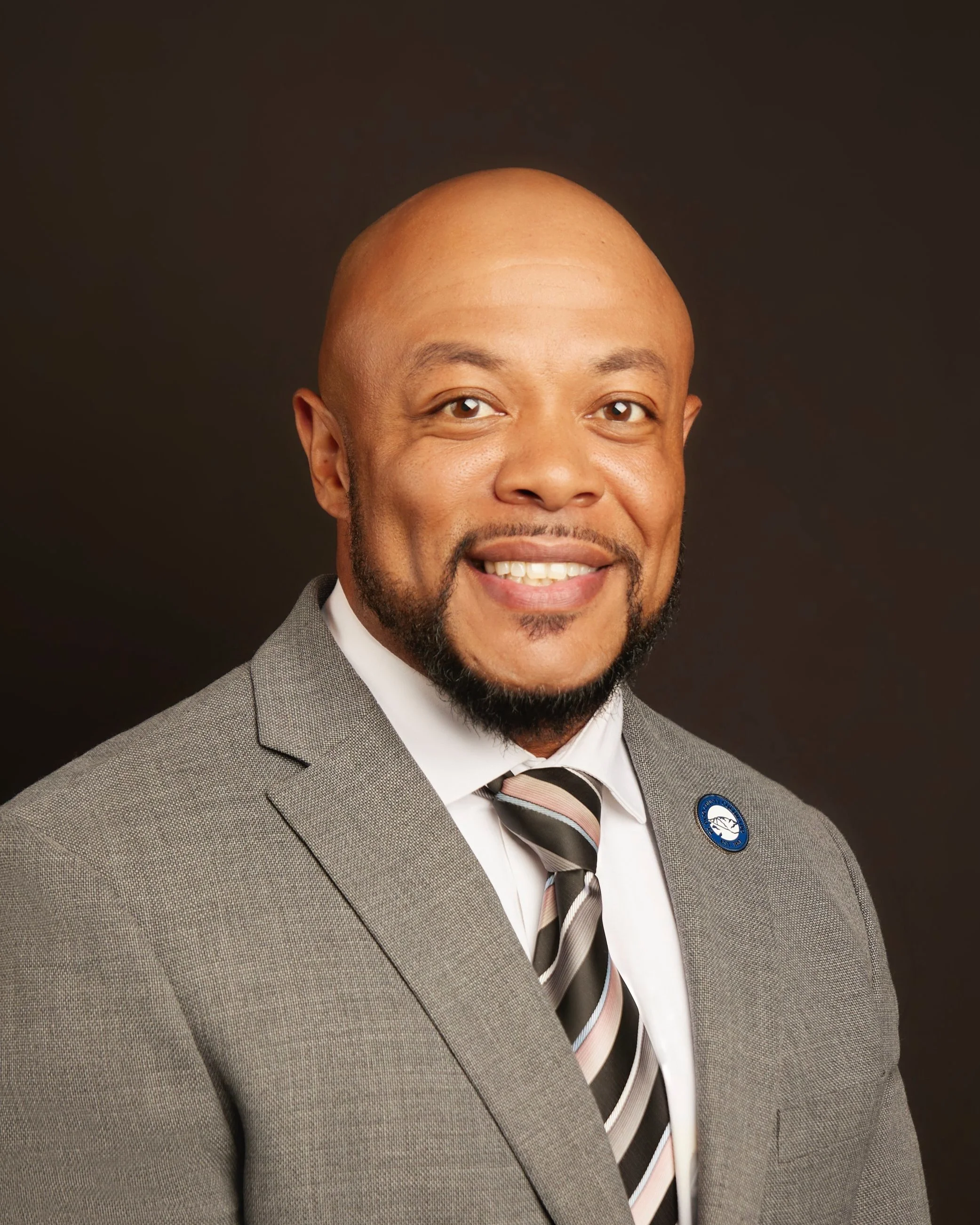 A smiling man with a bald head and a short beard wears a gray suit, white shirt, and striped tie, posing against a dark background.