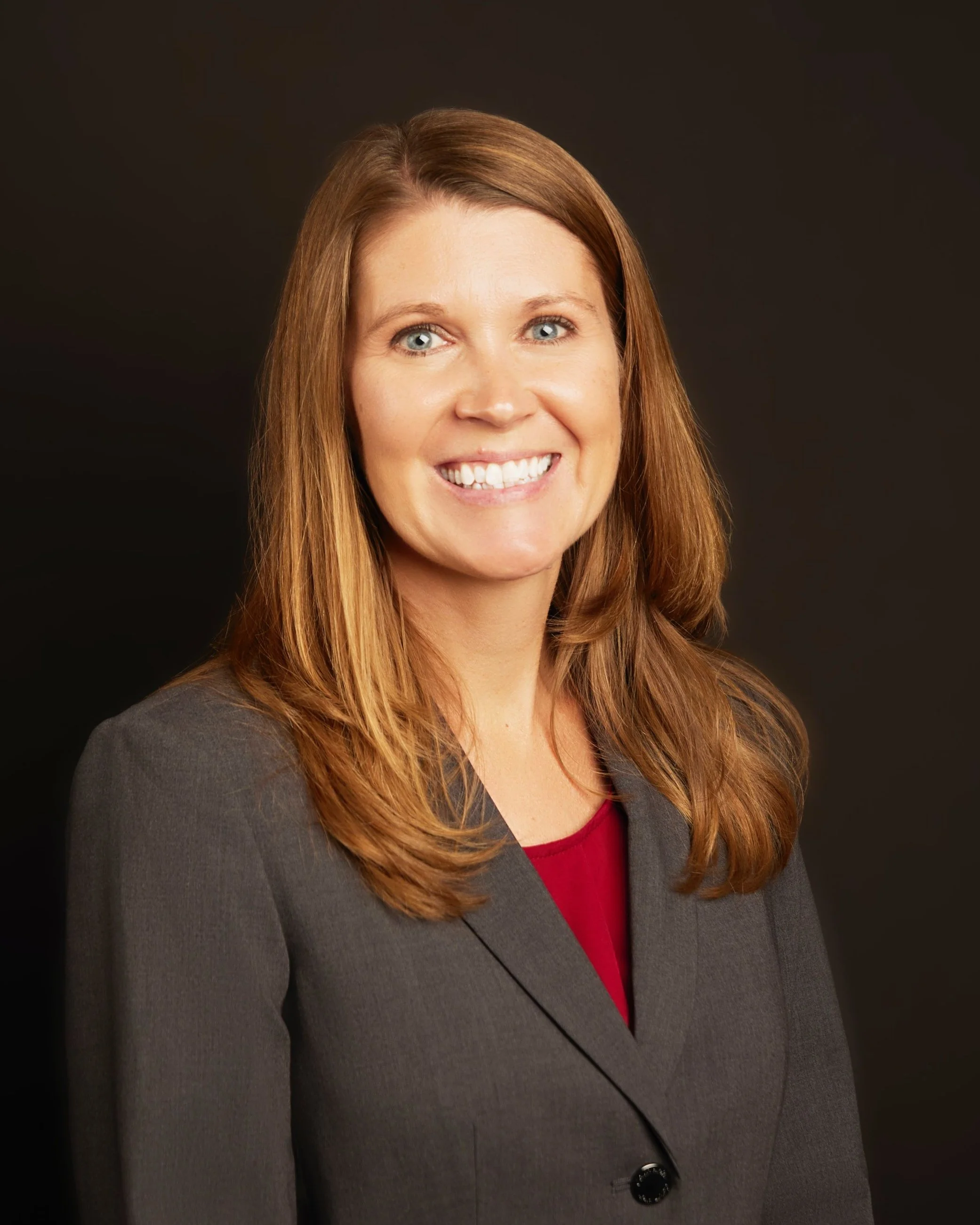 A smiling woman with long brown hair wears a gray blazer over a red top, posing against a dark background.