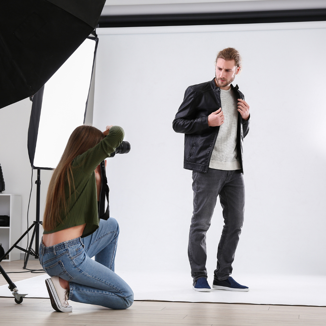 A photographer kneels to capture a shot of a man posing in a leather jacket and casual outfit in a professional studio setting.