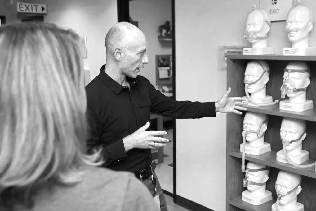 A man is explaining something to a woman in front of a shelf with mannequin heads wearing various medical headgear.