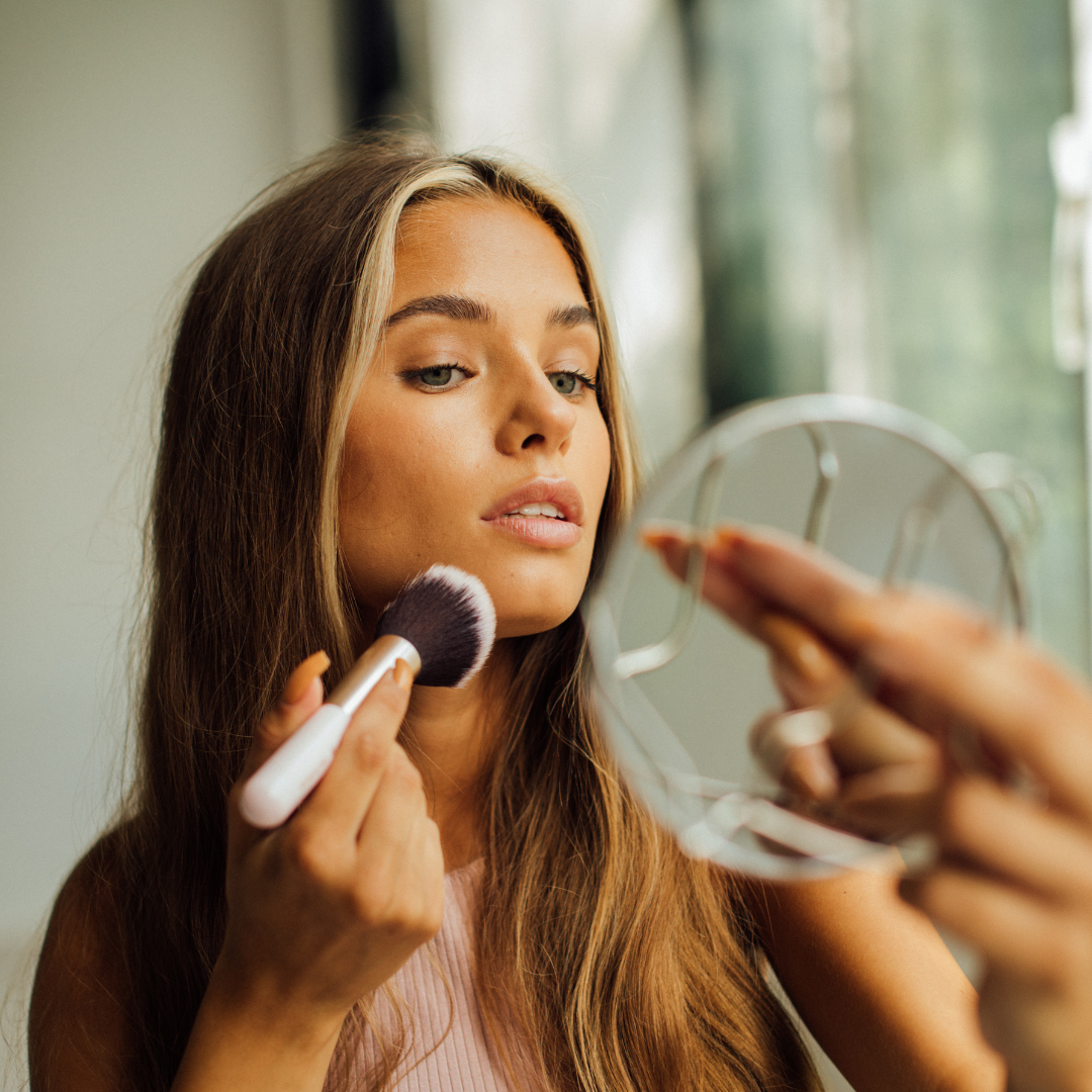 A woman with long hair applies makeup using a brush while looking into a small mirror.