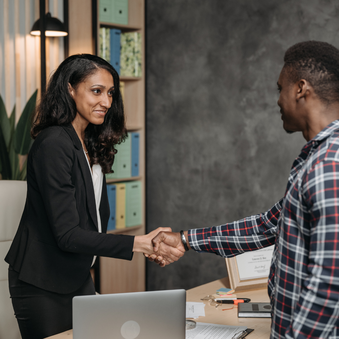 A woman in a black suit smiles and shakes hands with a man in a plaid shirt across a desk in an office.