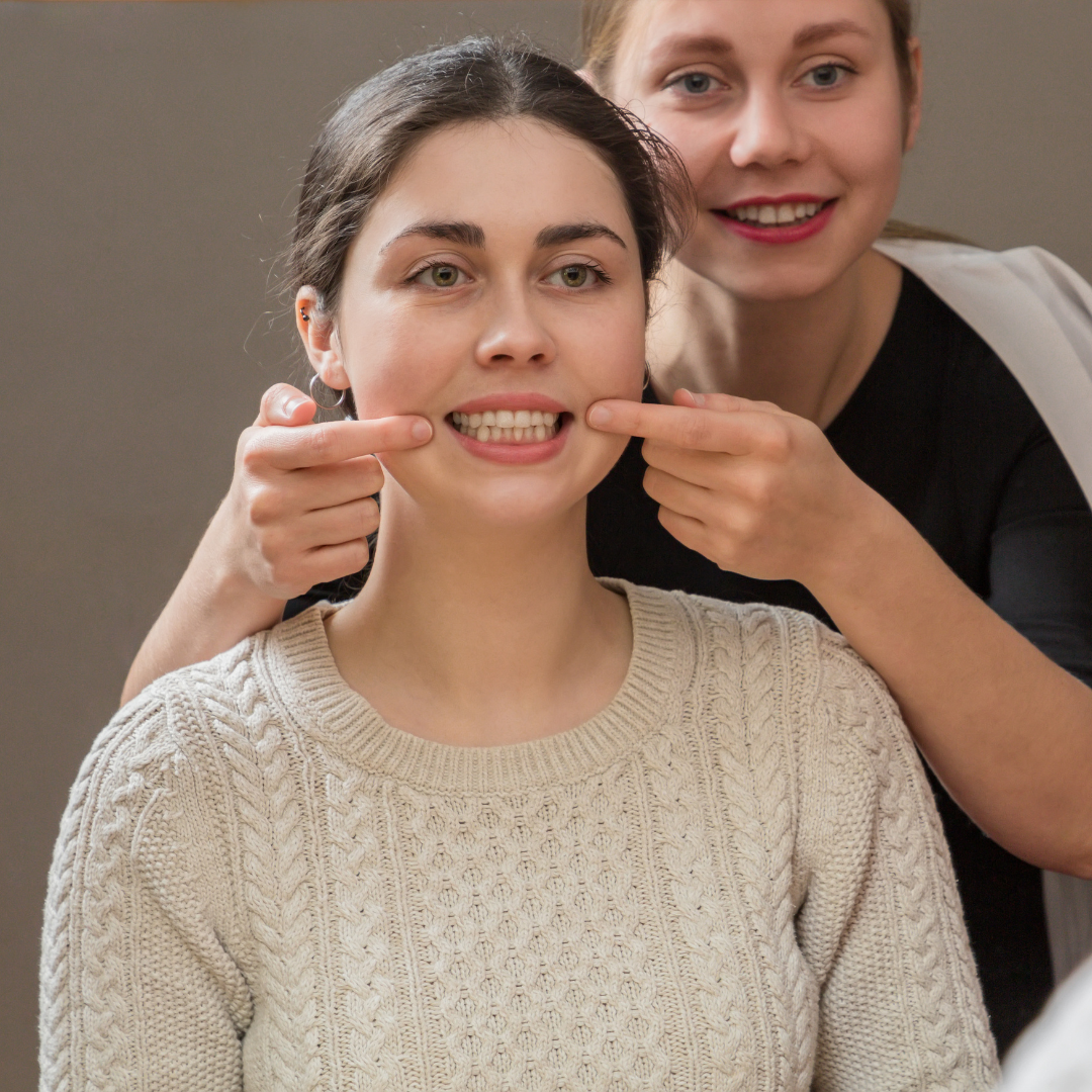 A woman in a sweater smiles as another woman gently adjusts her facial expression, possibly preparing for a photo.